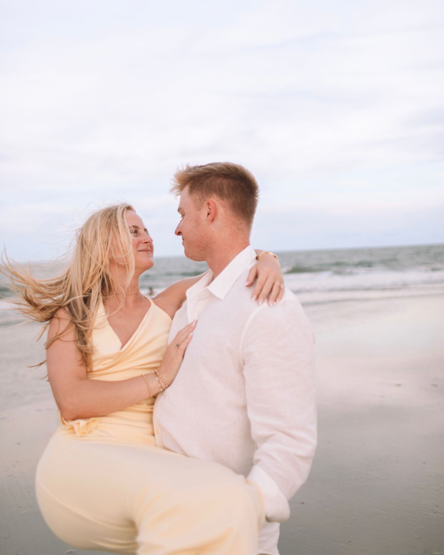 A man carries a woman in a yellow dress on a breezy beach at sunset.
