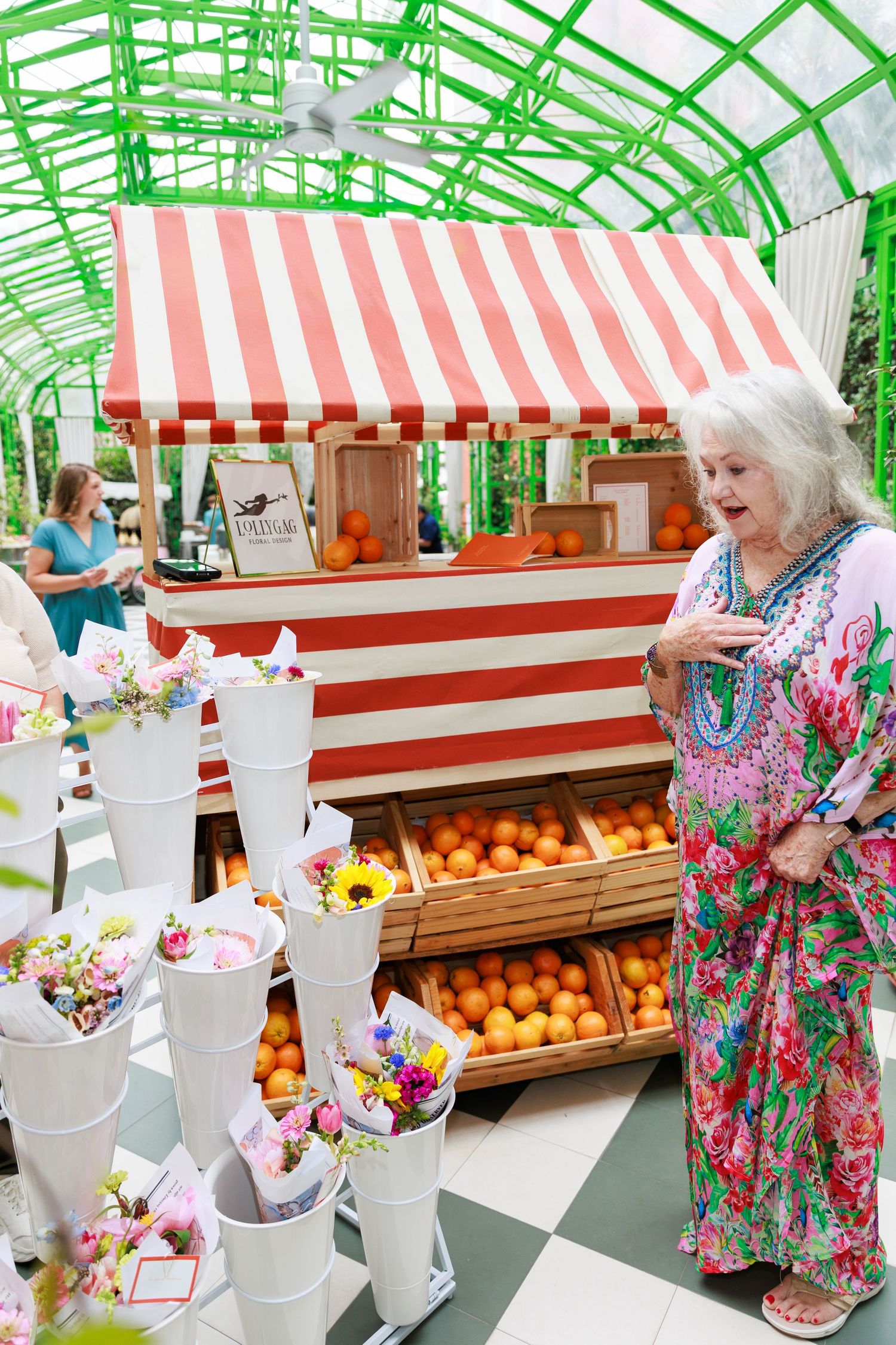 Elderly woman in floral dress browsing flower bouquets at a red and white striped market stall with fresh oranges.
