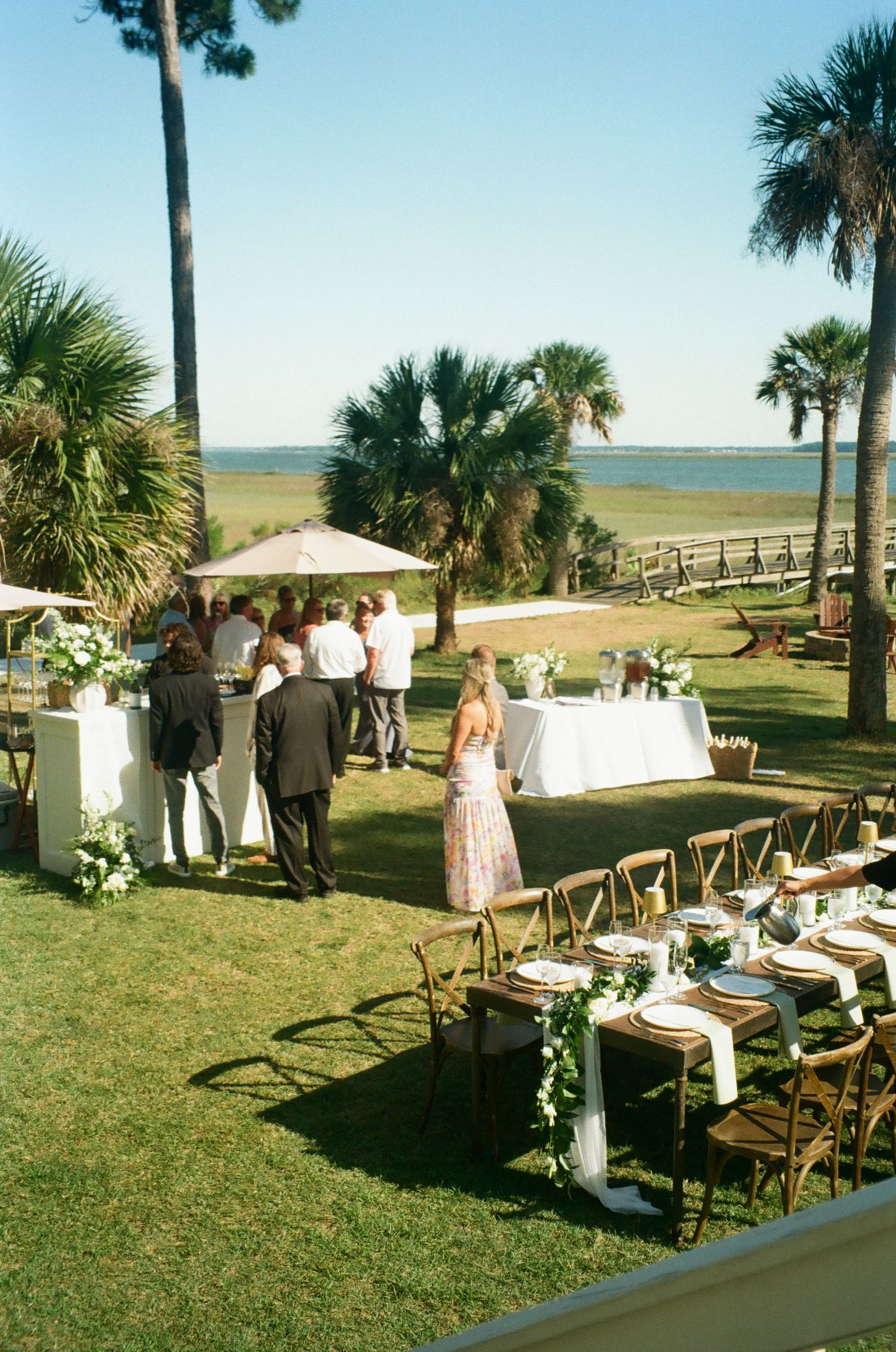 Outdoor waterfront wedding reception with long wooden tables, cross-back chairs, palm trees, and guests mingling by the bar.