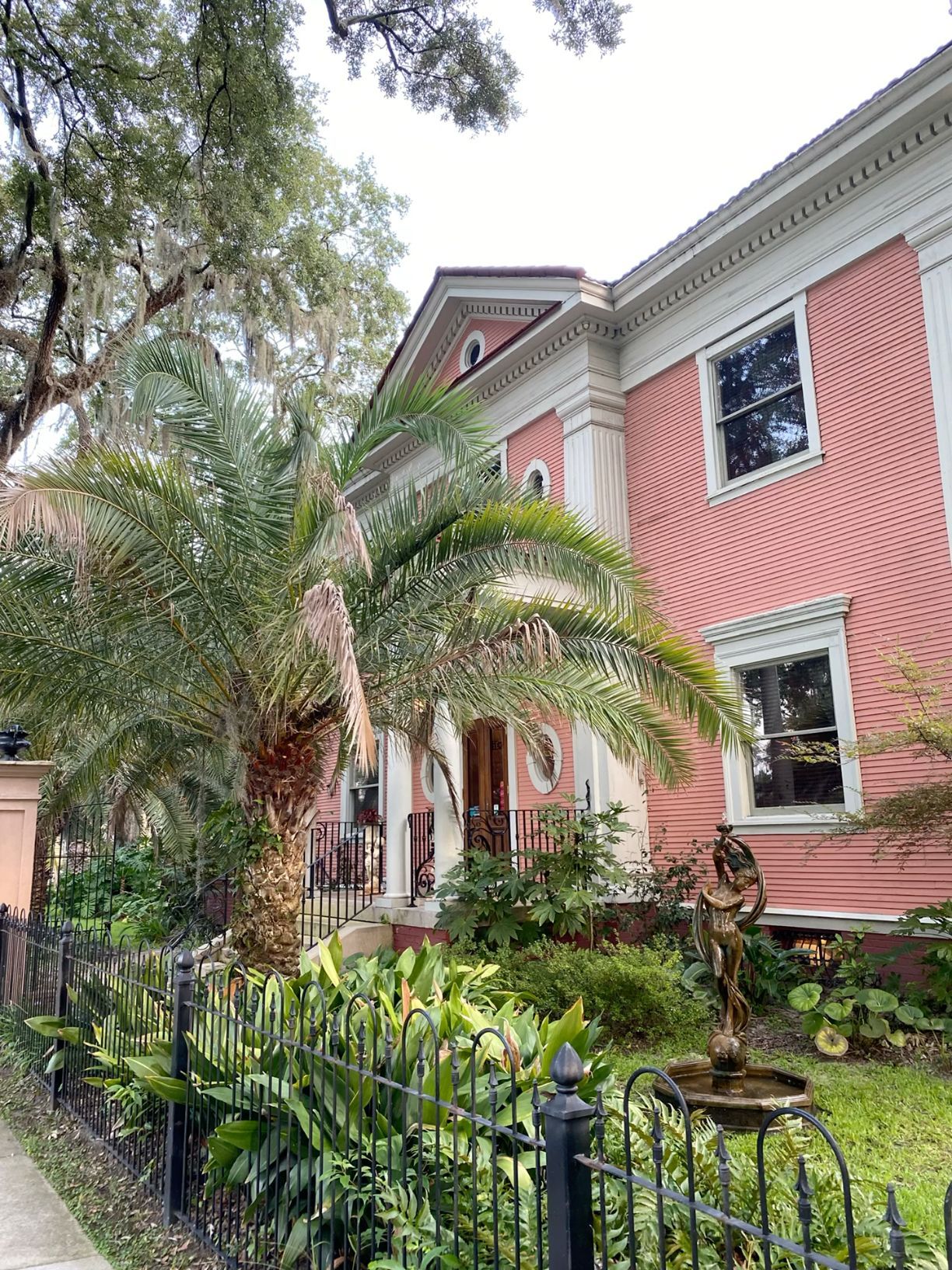 Pink historic mansion with palm trees and lush garden behind iron fence.