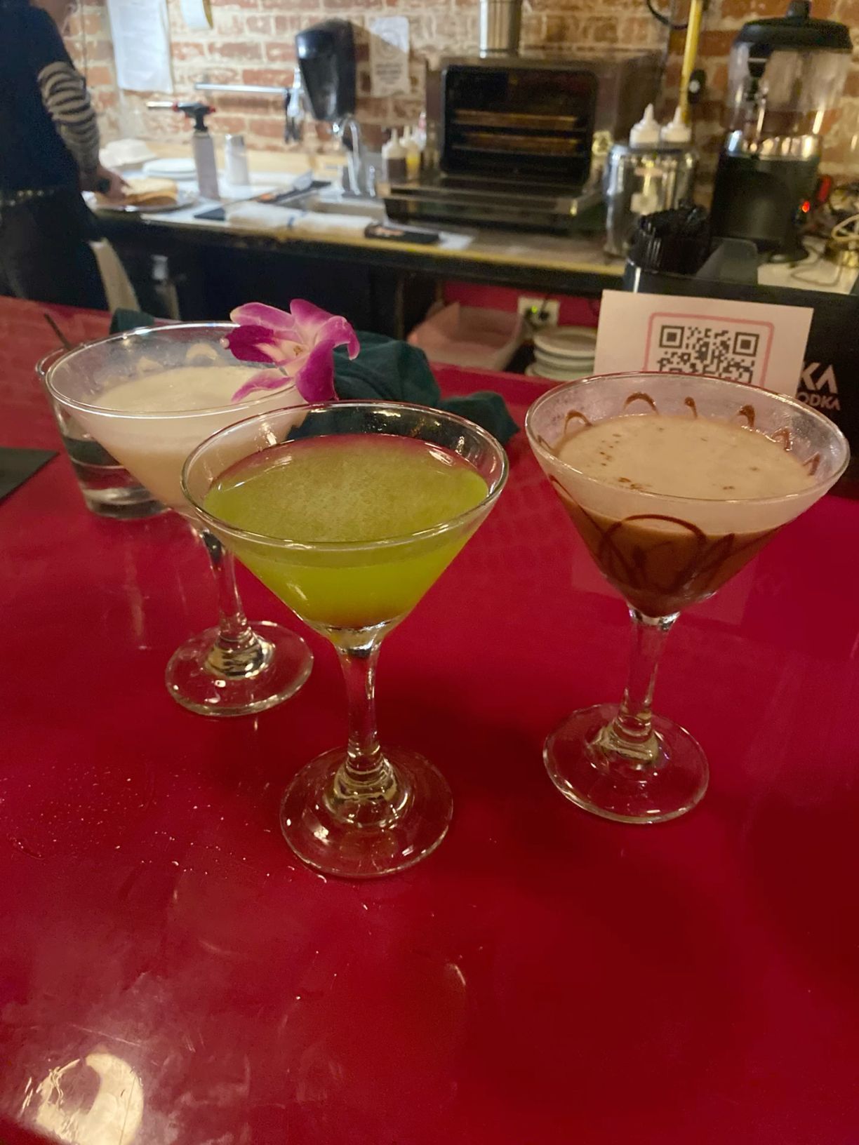 Three decorative cocktails on red bar including green martini and drinks with floral garnish.