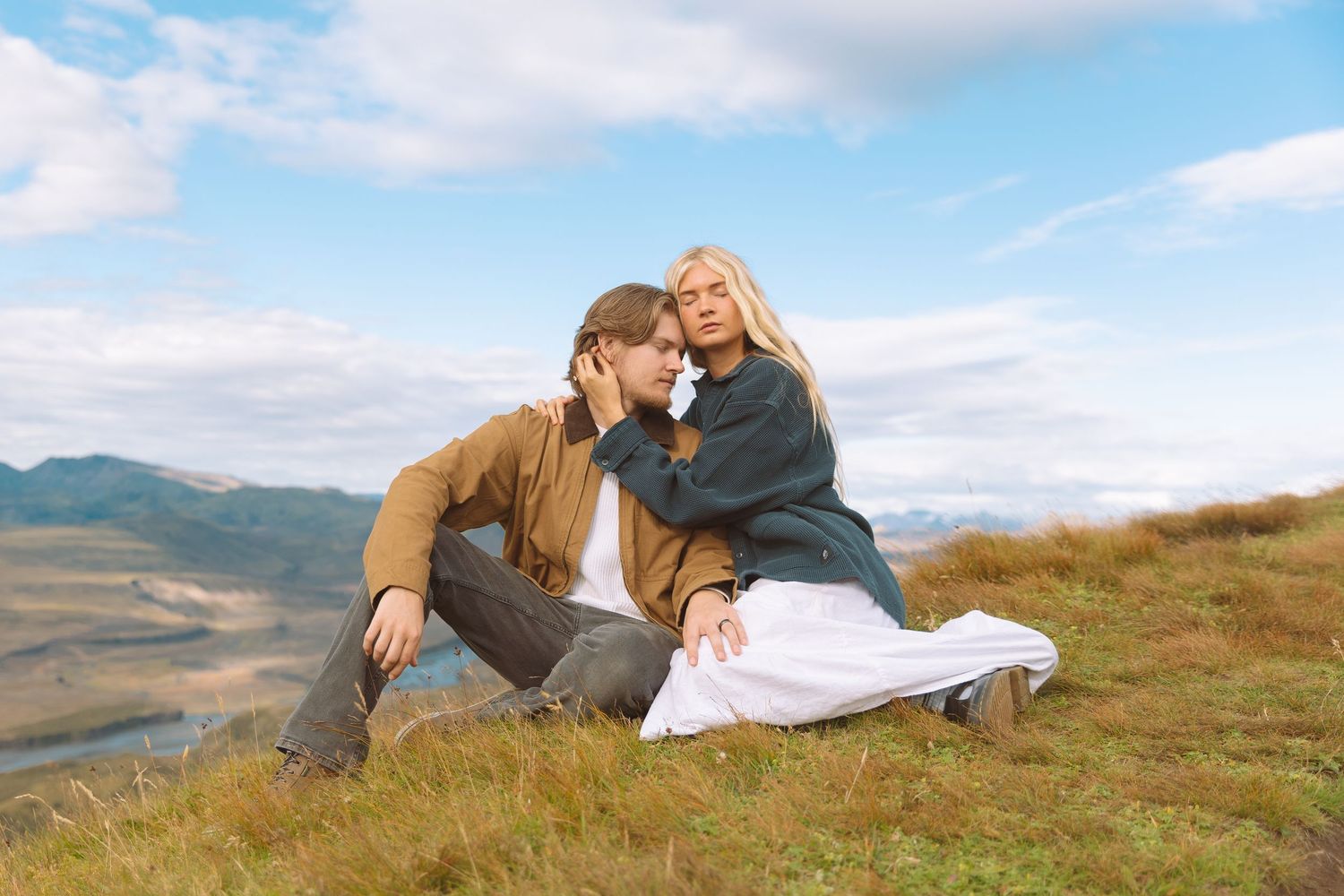Couple sitting on a grassy hillside with mountain landscape backdrop.