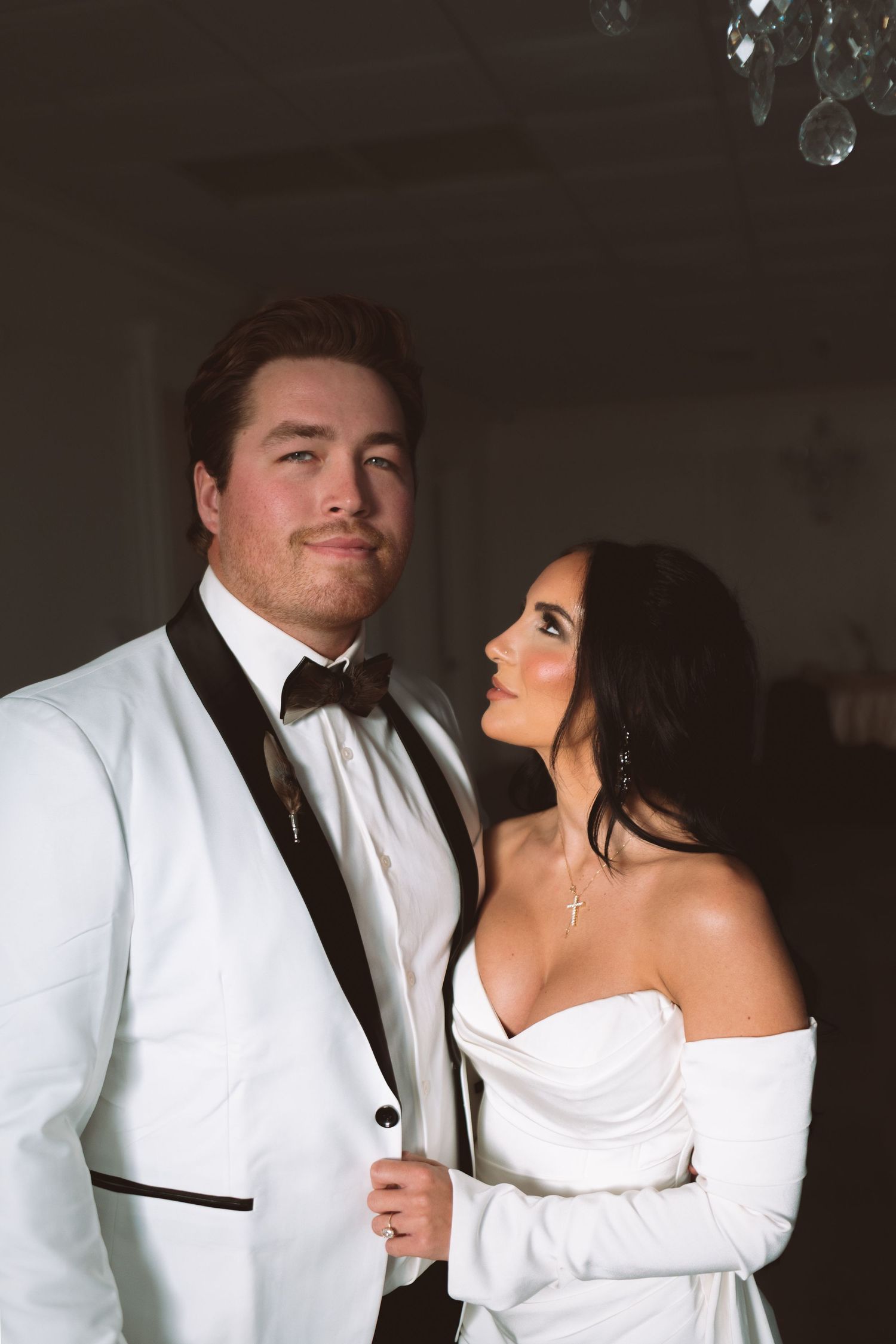 Bride in white off-shoulder gown gazes up at groom in white tuxedo with black lapels in a dramatic indoor setting.