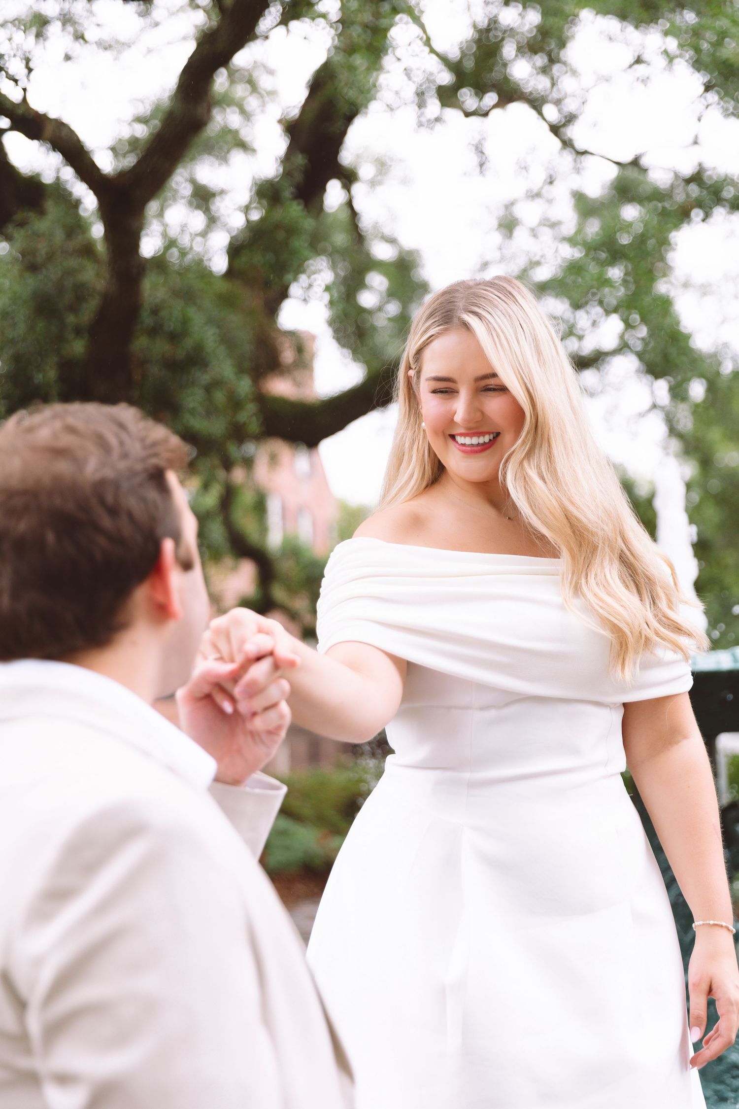 Smiling blonde bride in off-shoulder white gown holds groom's hand outdoors under lush green trees.