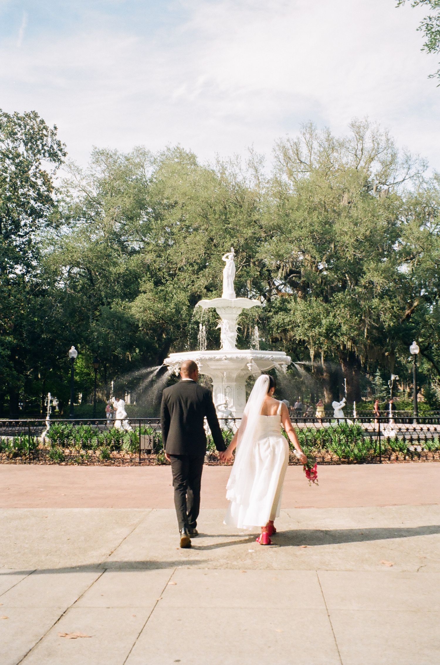 Newlyweds walking hand-in-hand toward iconic white fountain in Forsyth Park, Savannah, Georgia.