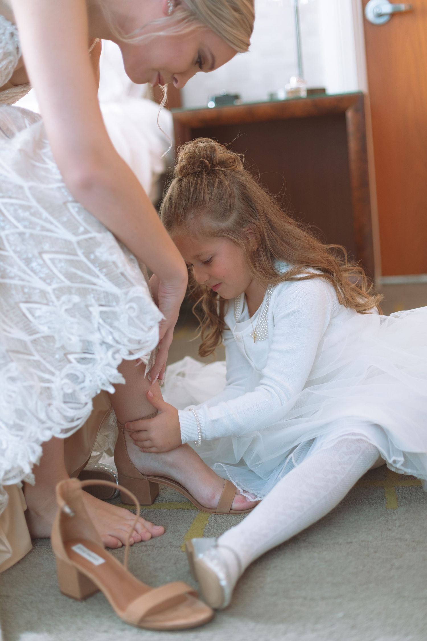 Flower girl in white dress helps bride put on nude block heel wedding shoes before ceremony.