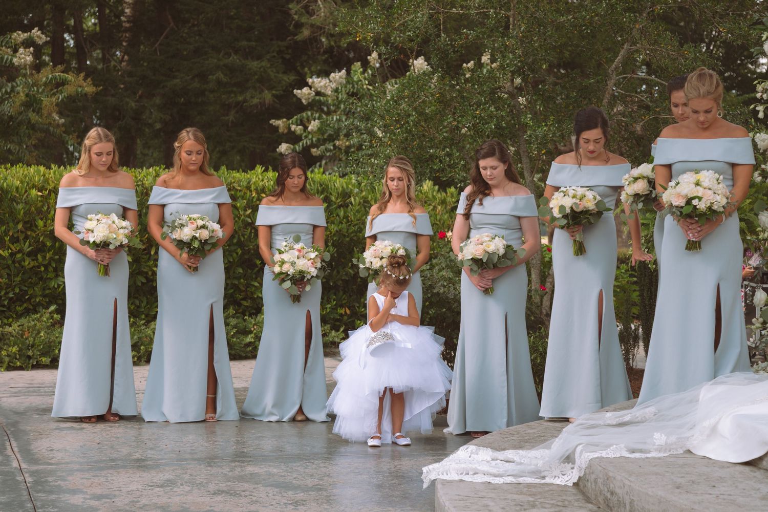 Bridesmaids in light blue off-shoulder gowns holding white bouquets with flower girl at outdoor ceremony.