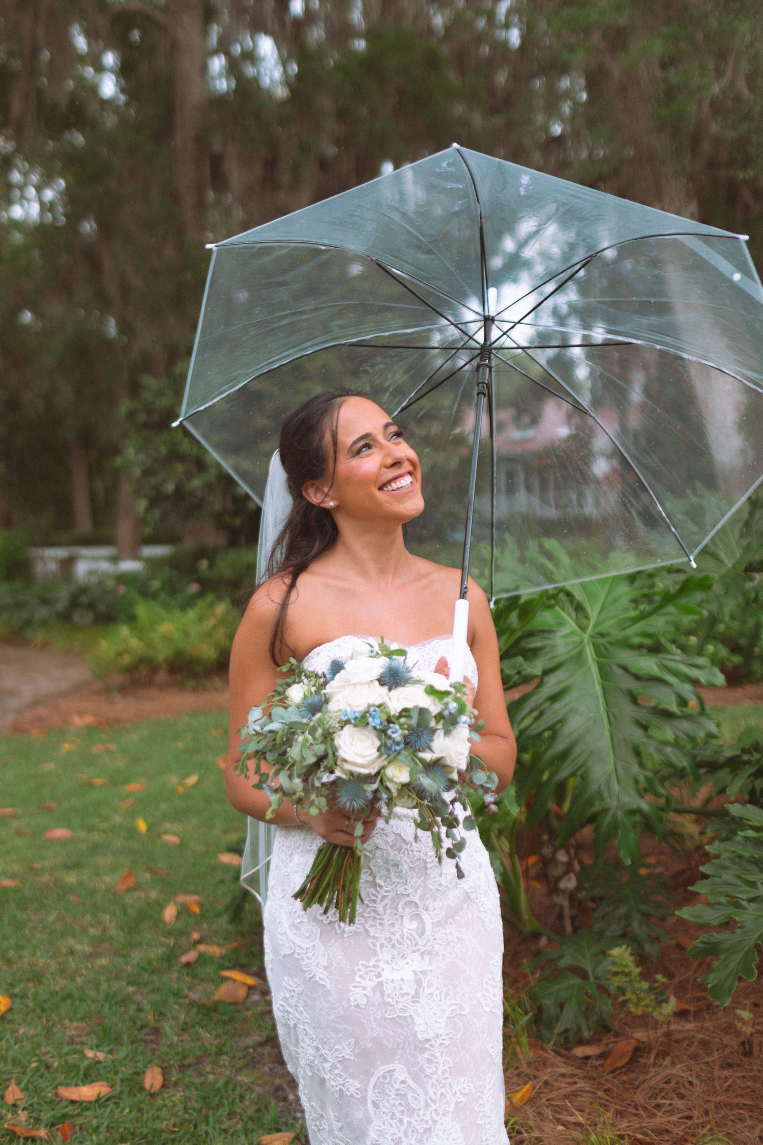 Smiling bride in white lace gown holds blue and white bouquet under clear umbrella in lush garden setting.