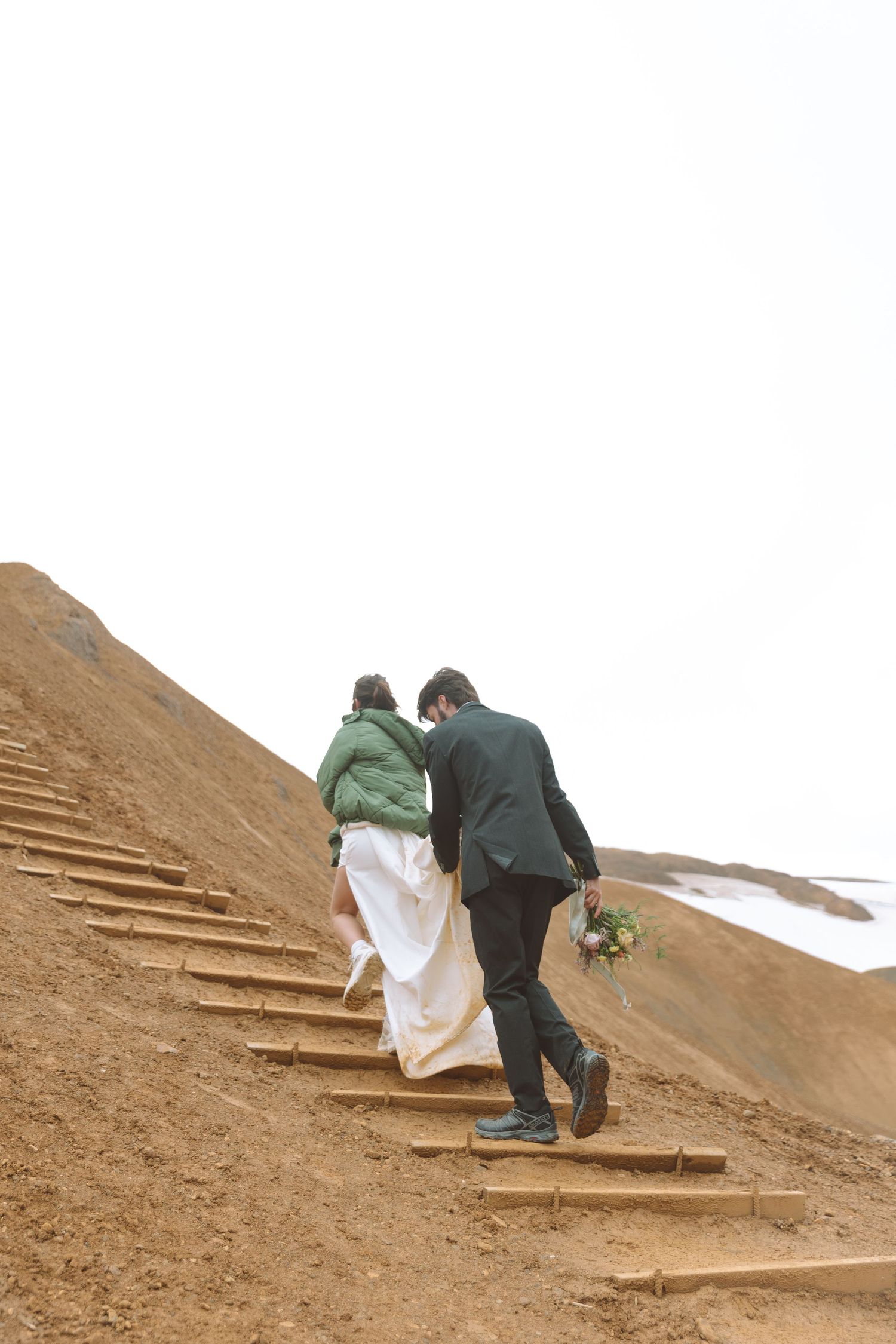 Wedding couple climbs rustic wooden stairs up Iceland's golden rhyolite mountains with bouquet in hand.