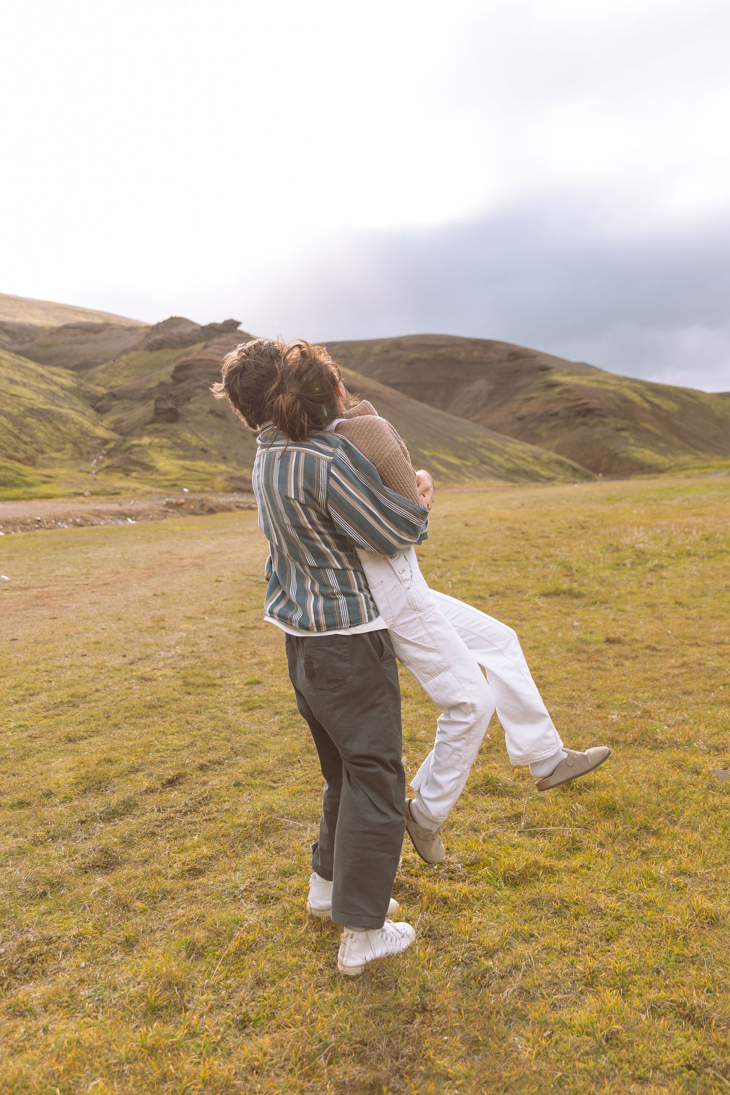 Playful couple hugging and lifting each other in a scenic green mountainous landscape.