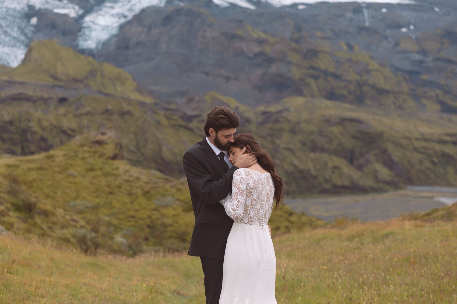 Newlywed couple shares a quiet embrace in a vast green valley with dramatic glacier mountains behind.