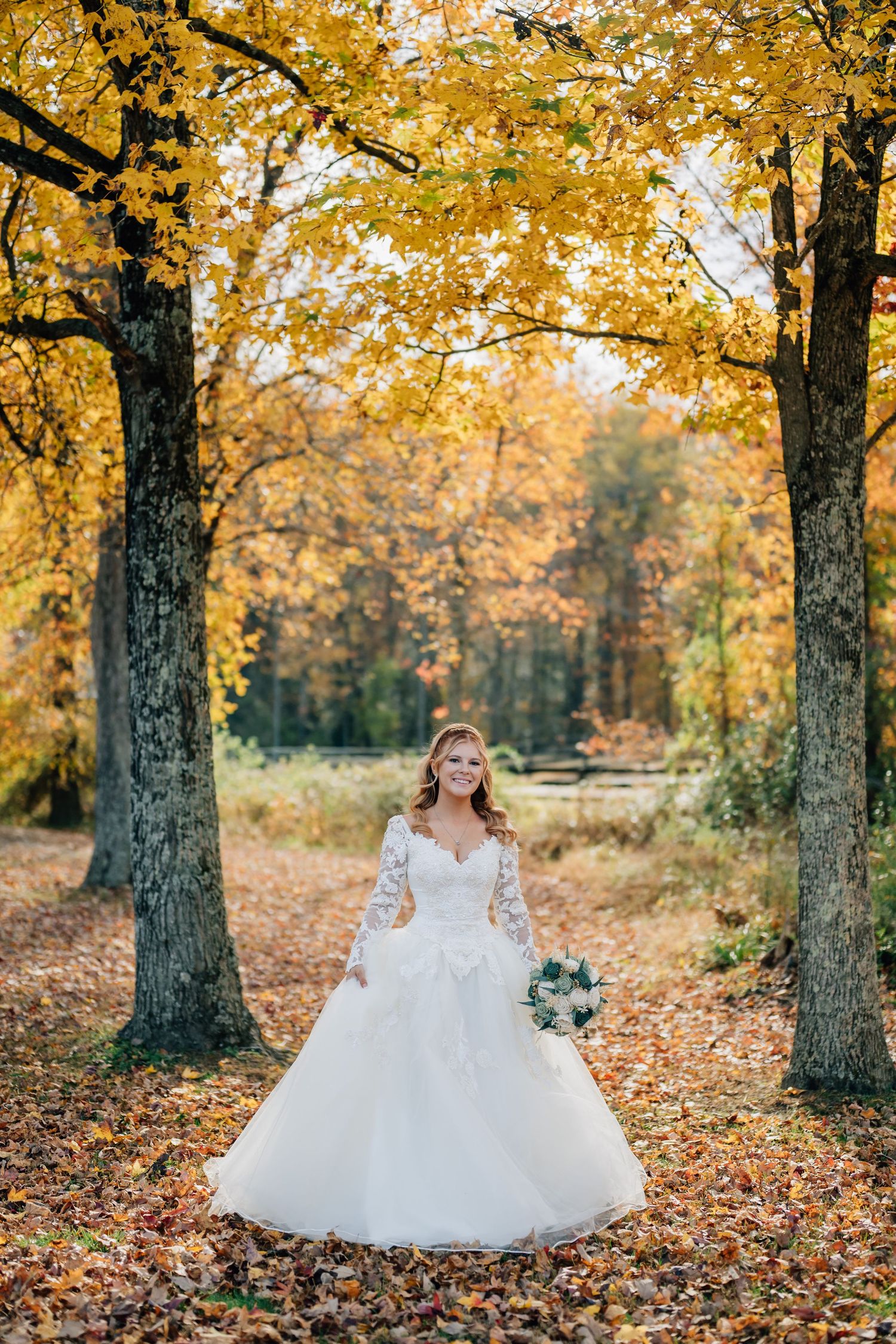 Bride in wedding dress standing beneath golden autumn trees with fallen leaves at Arbor Haven Weddings in Fredericksburg, VA.