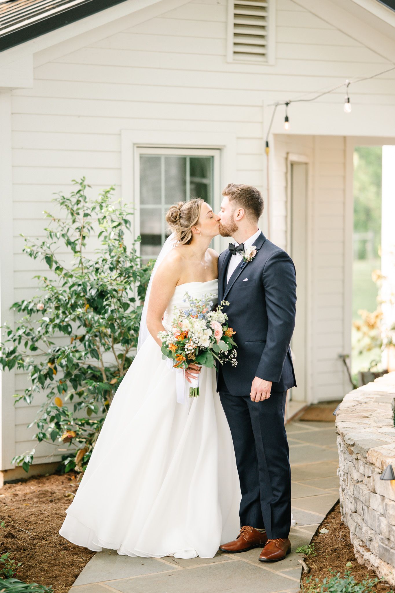 A wedding couple shares intimate moments outside a white farmhouse building.