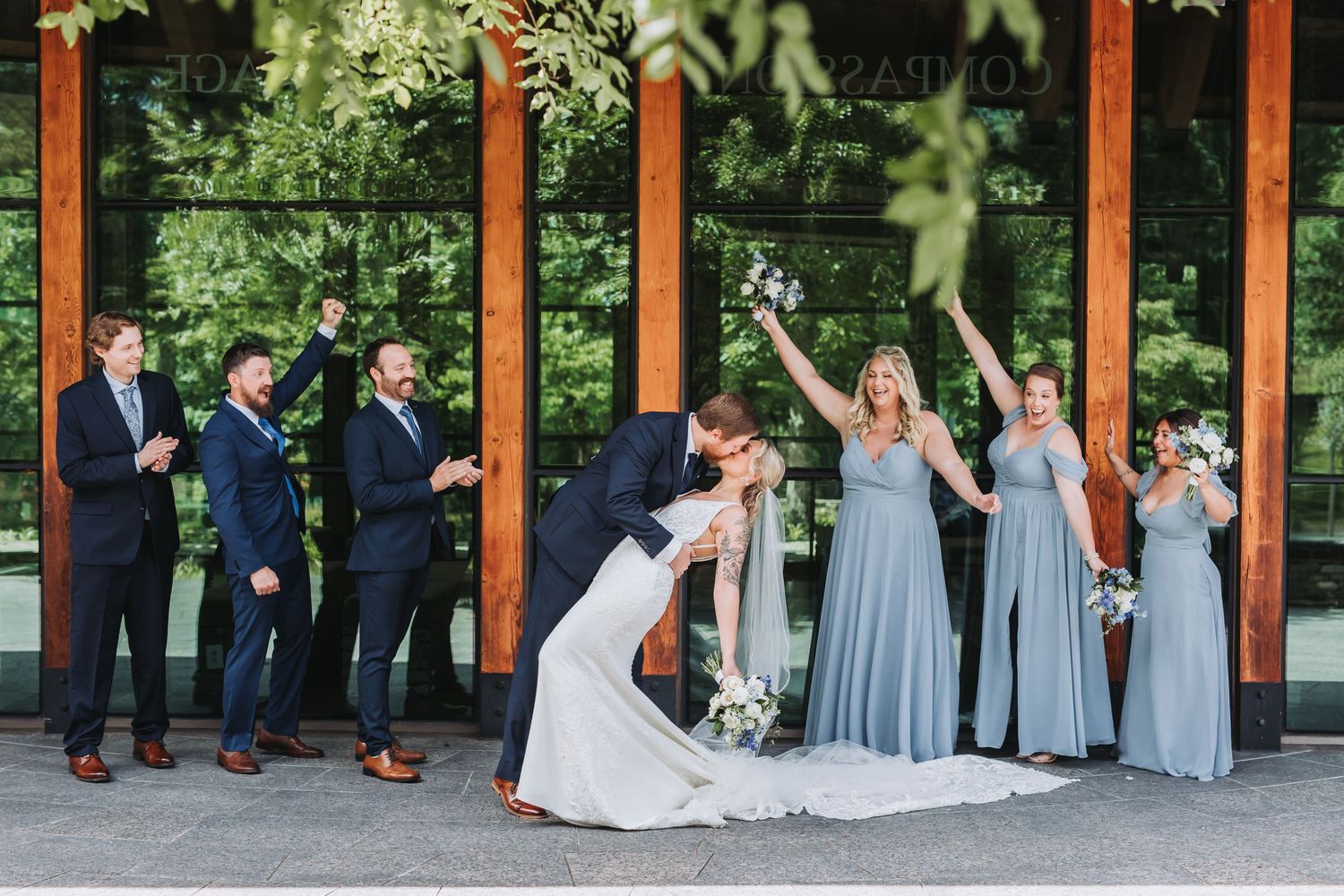 Wedding party poses under wooden pavilion with bridesmaids in grey dresses.