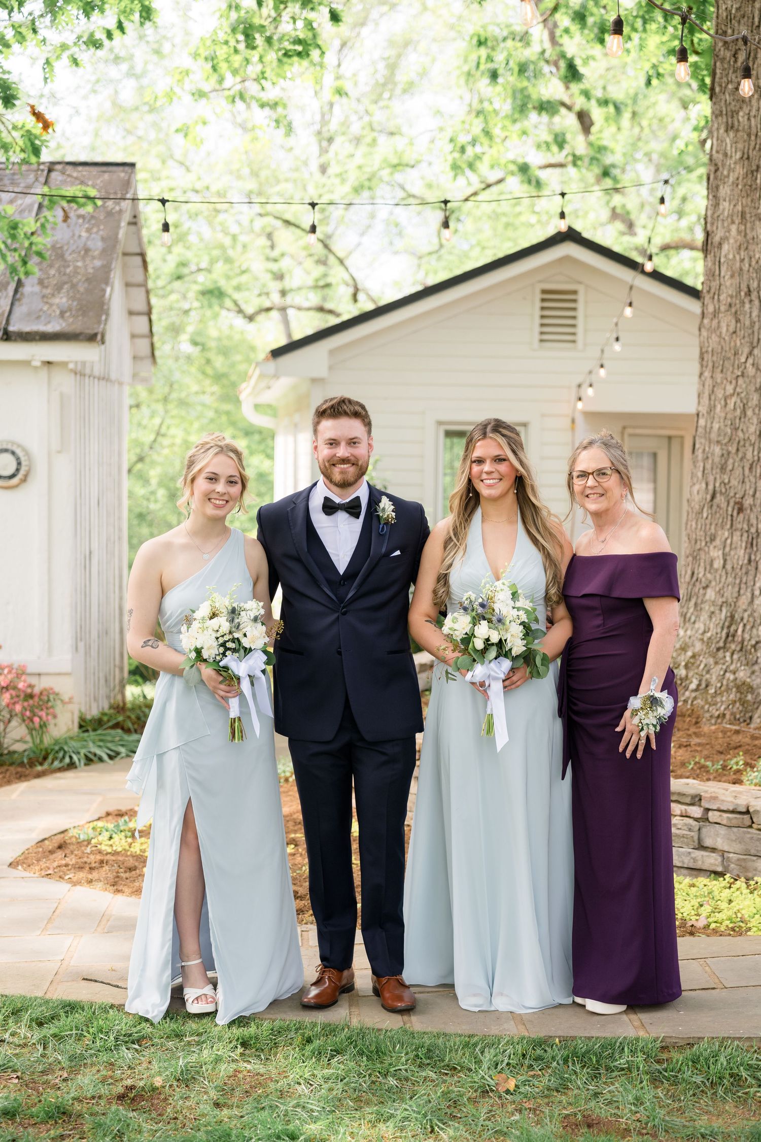 Wedding party in formal attire holding bouquets poses together on rustic patio.
