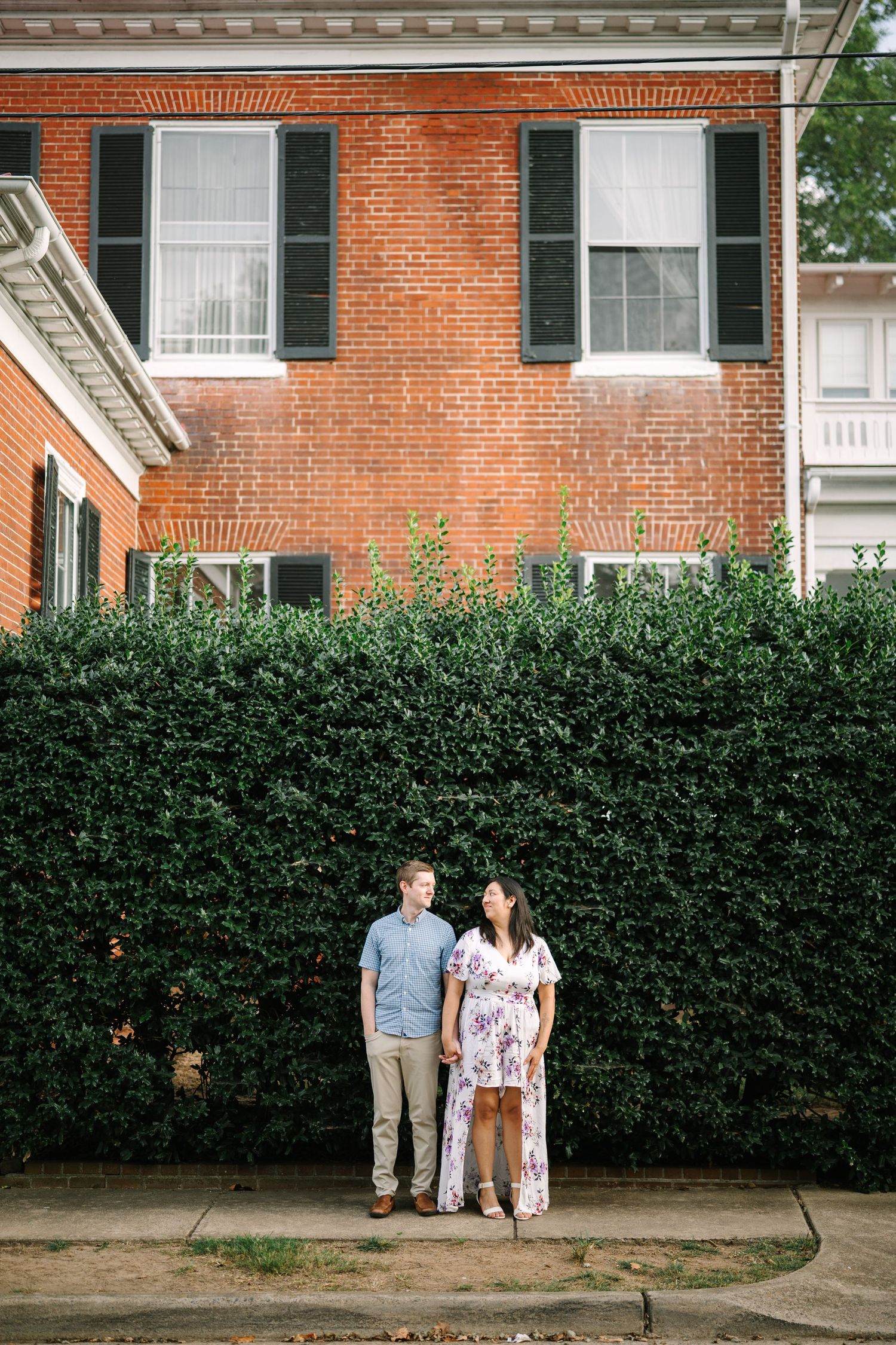 Couple standing against a brick wall with green hedge in a series of romantic outdoor photos.