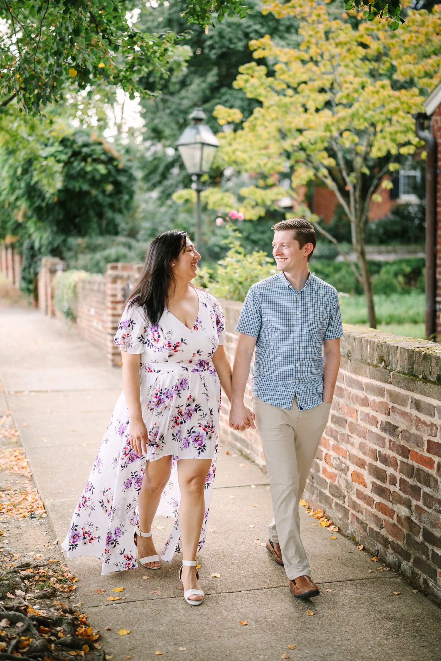 Couple walking hand in hand along brick wall path under autumn trees.
