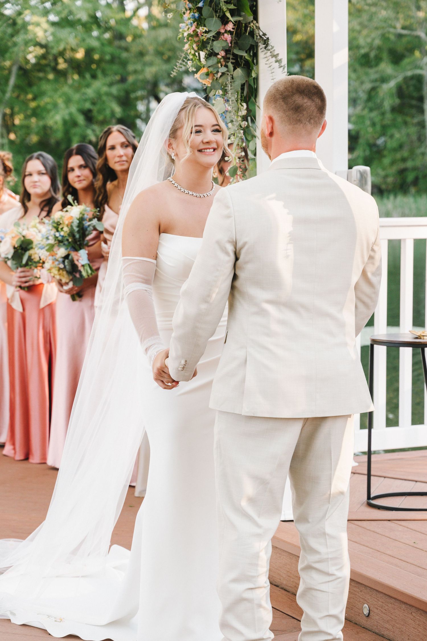 Wedding ceremony participants gather on a wooden deck surrounded by coral-colored dresses.