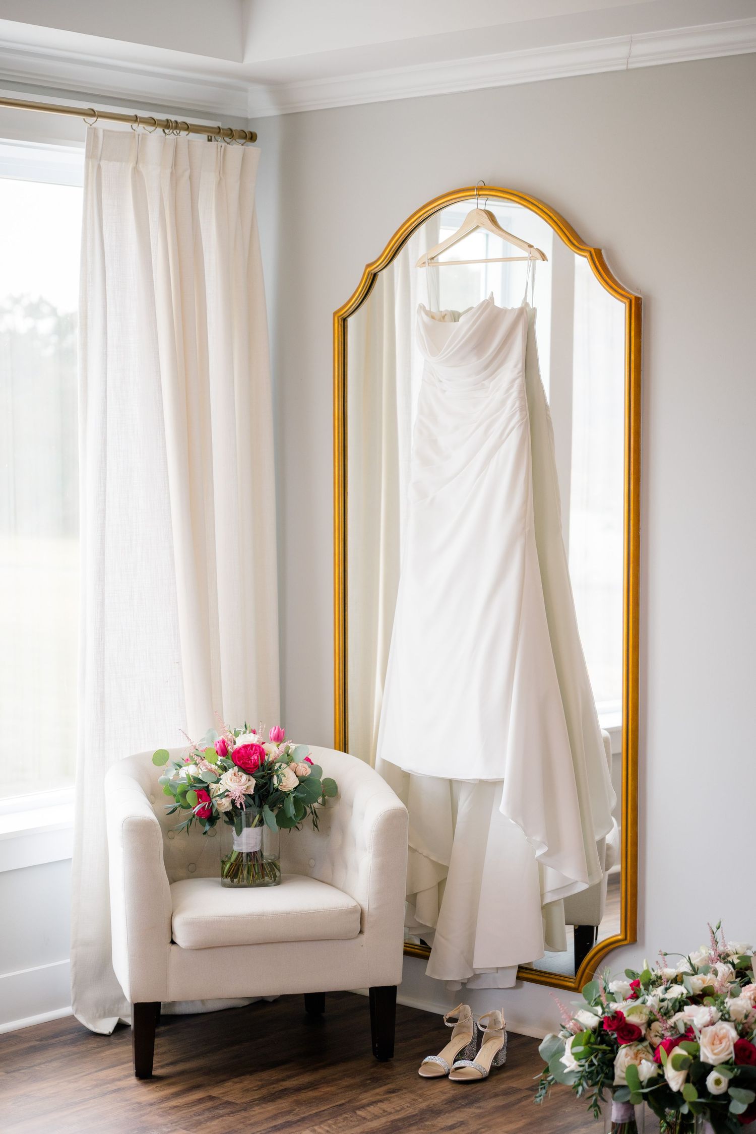Light-filled room with white curtains, gold-framed mirror, and pink floral arrangements on the floor.