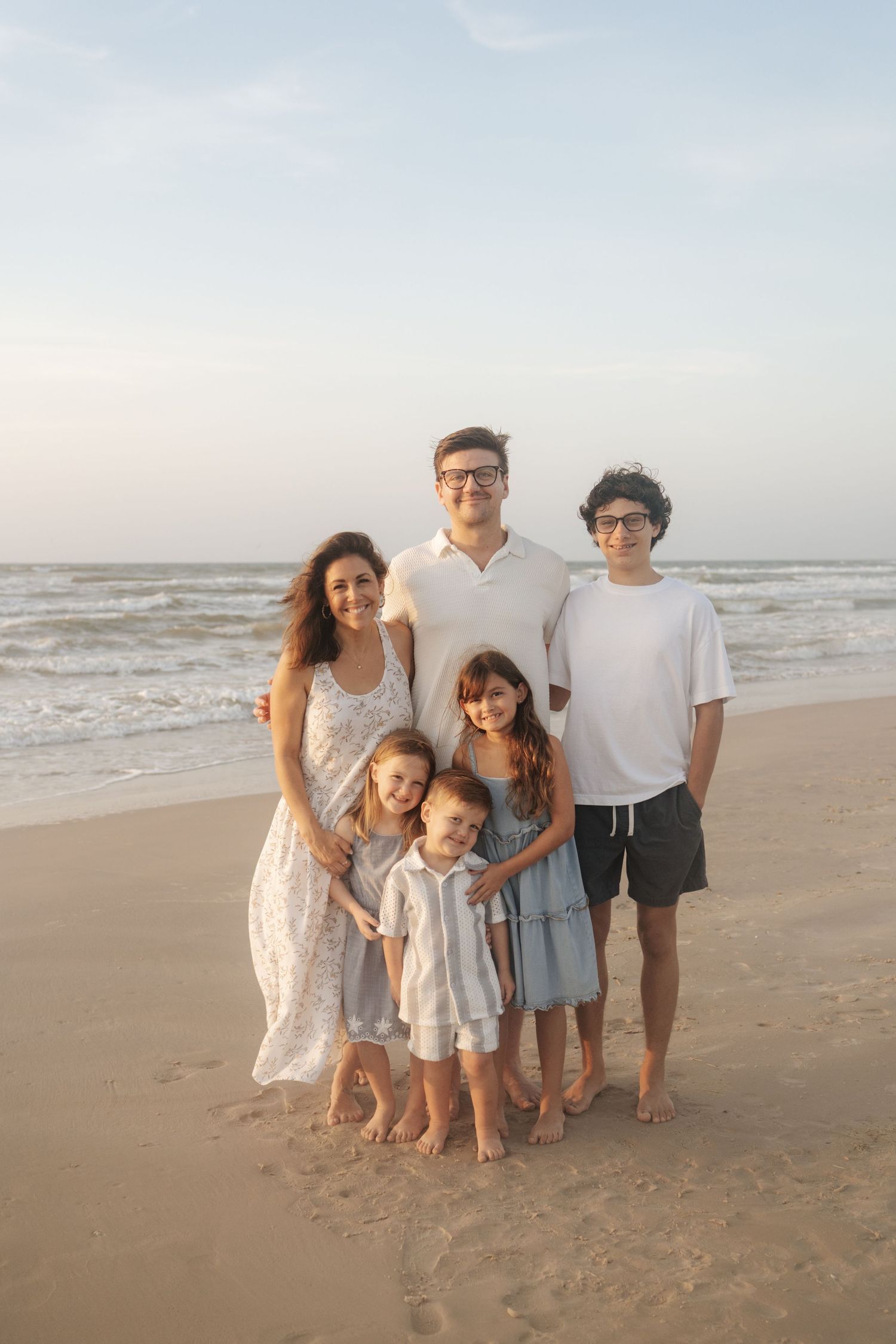 Family of five posing together on a sandy beach at sunset, with ocean waves in the background.