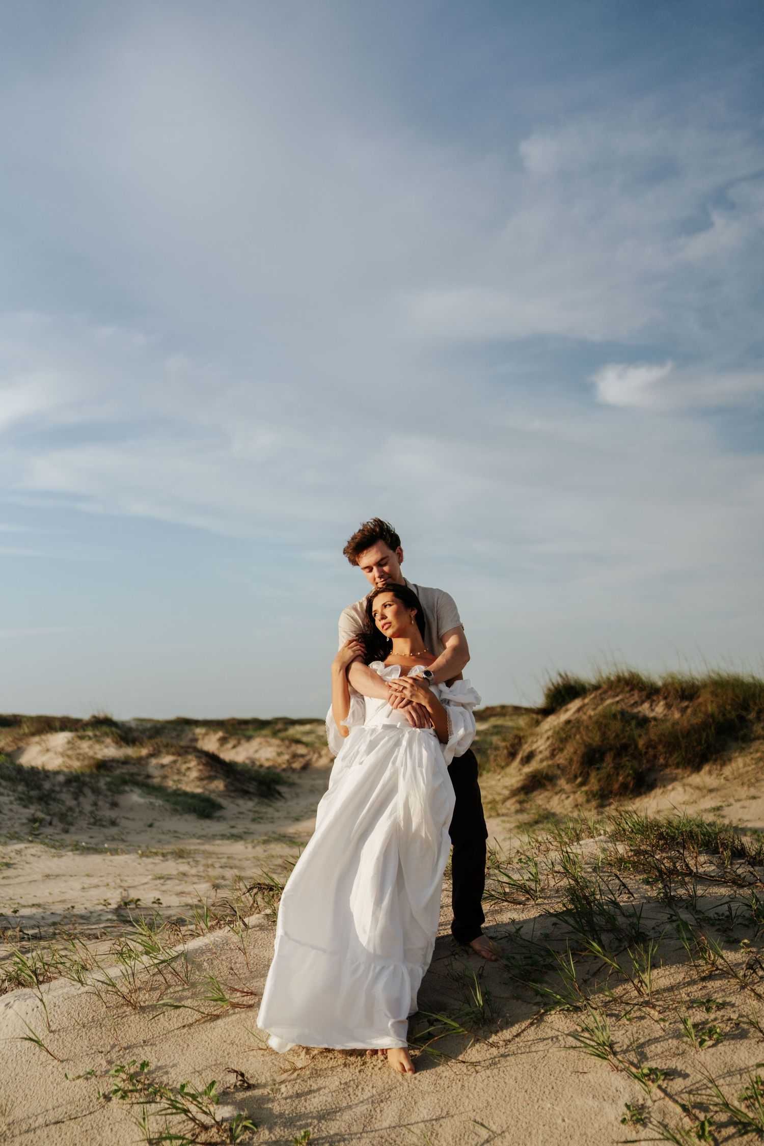 Couple embracing on sandy beach dunes, woman in white dress, man in white shirt, blue sky backdrop.
