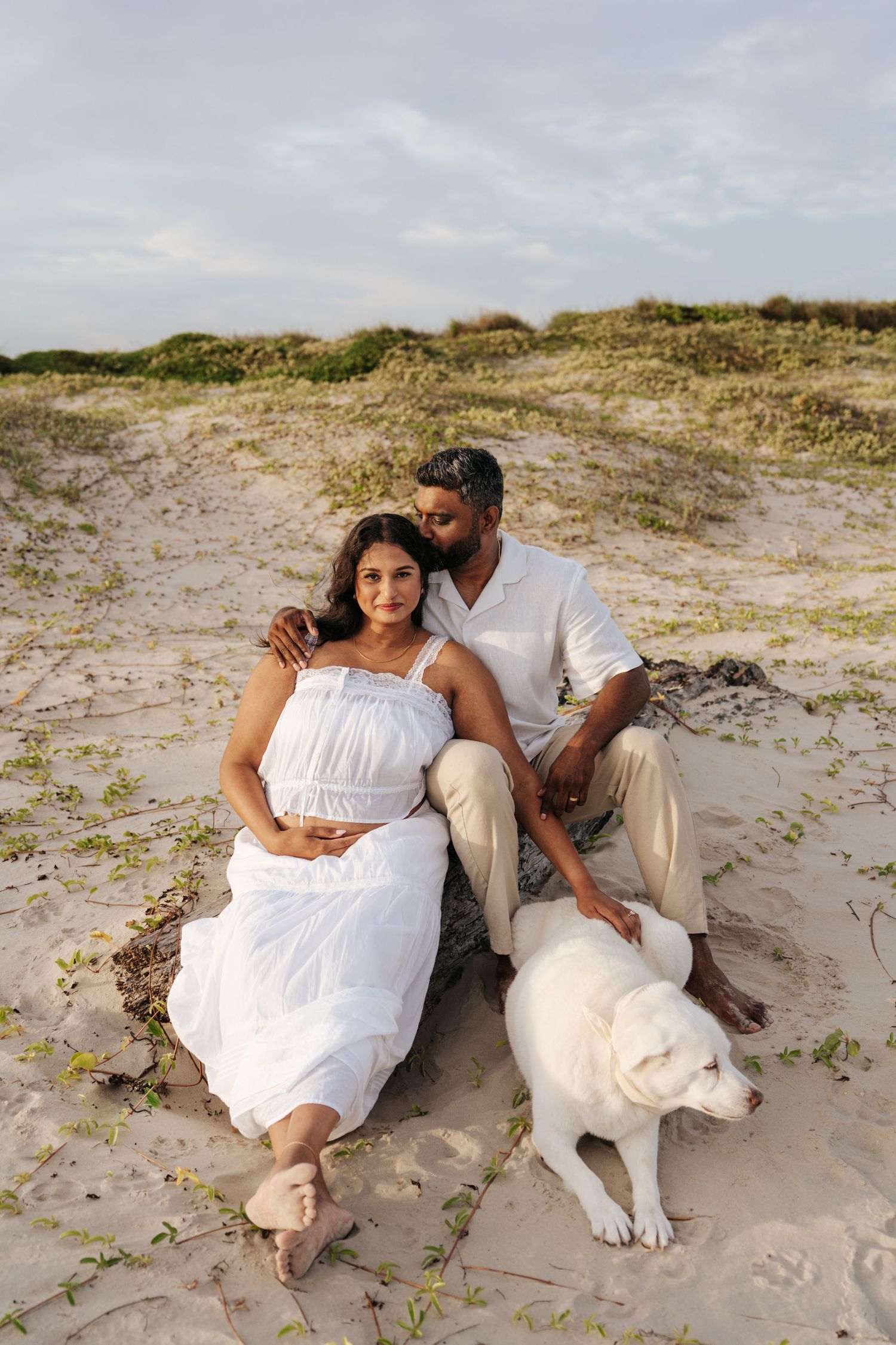 Couple in white outfits sitting on sandy beach dunes with a white Labrador dog during golden hour.
