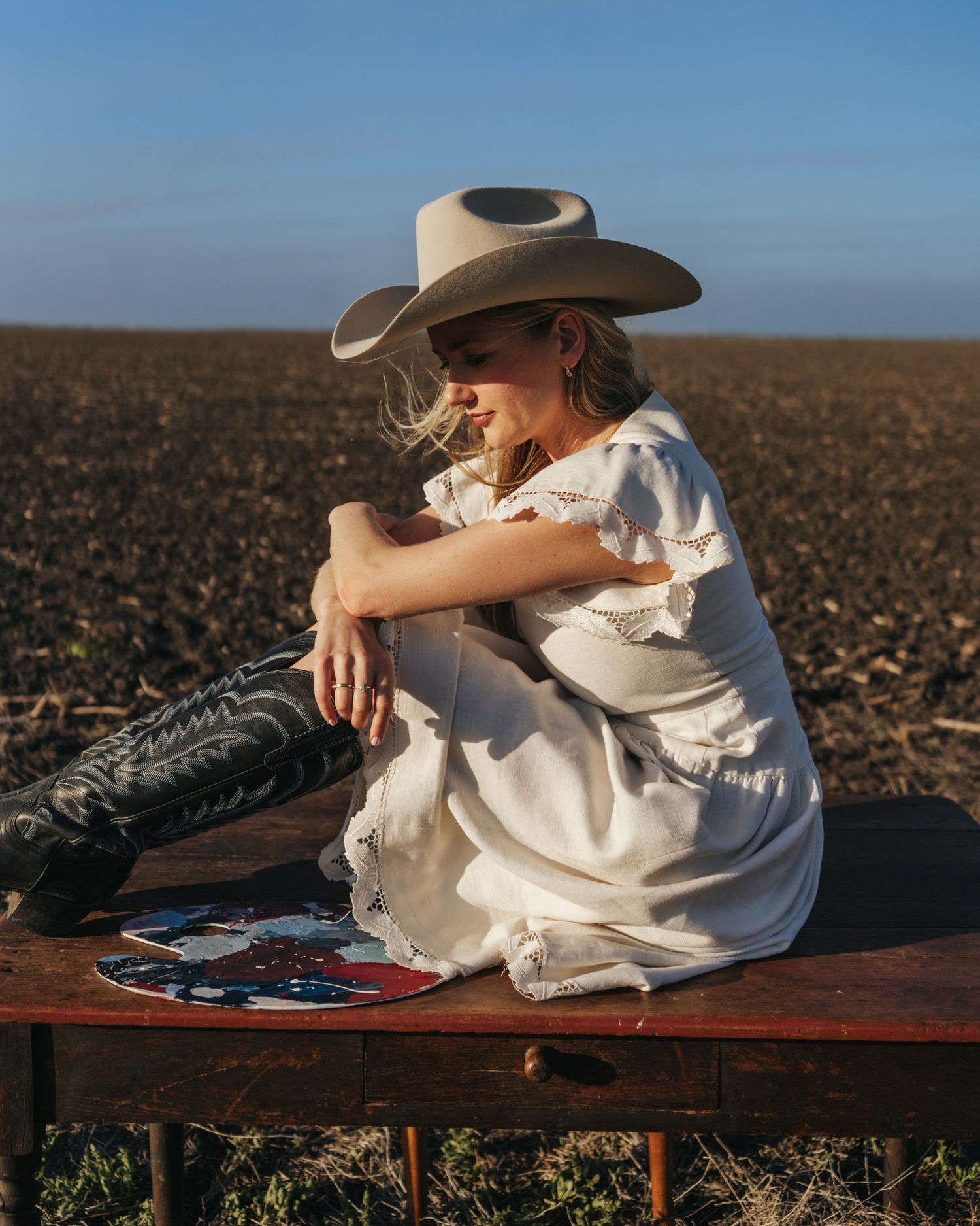 Woman in white dress, cowboy hat, and black boots sits on a table in a field holding a colorful fan.