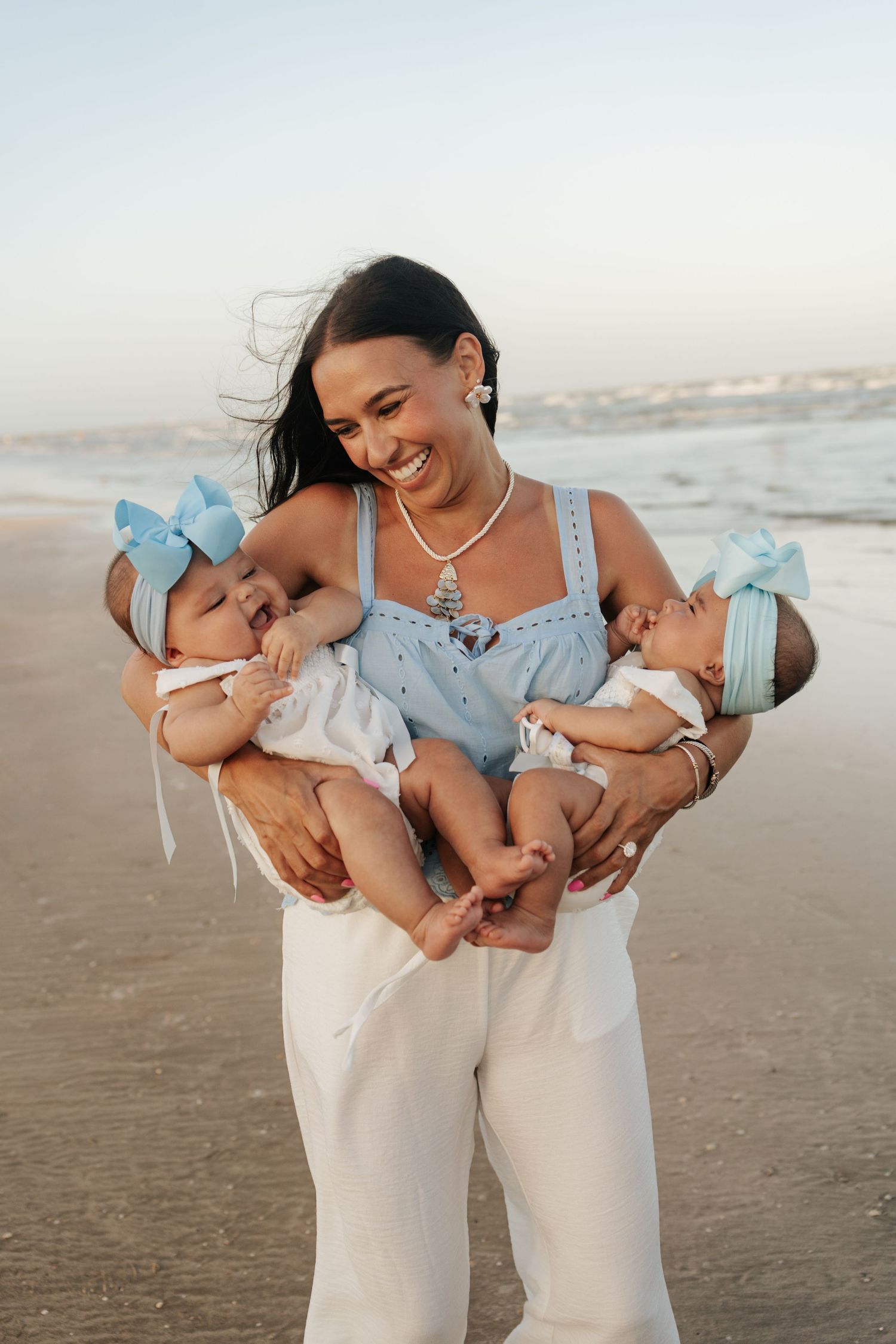 Smiling woman in blue top holds twin babies with blue bows on a sunny beach.