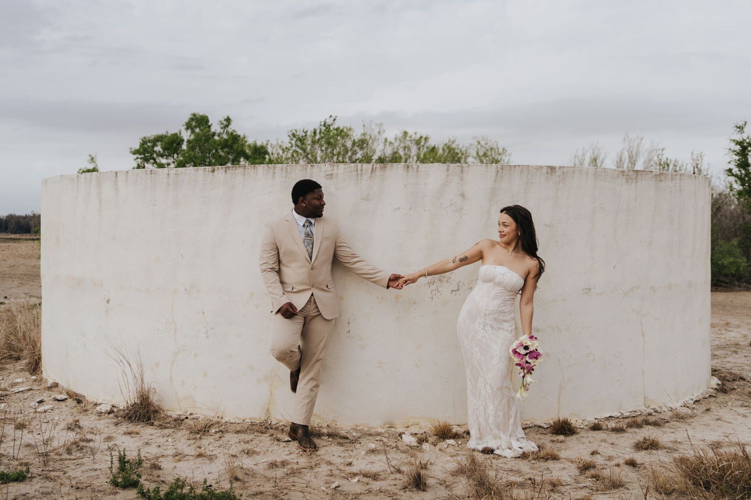 Interracial couple in wedding attire holding hands against a white concrete wall in an open field.
