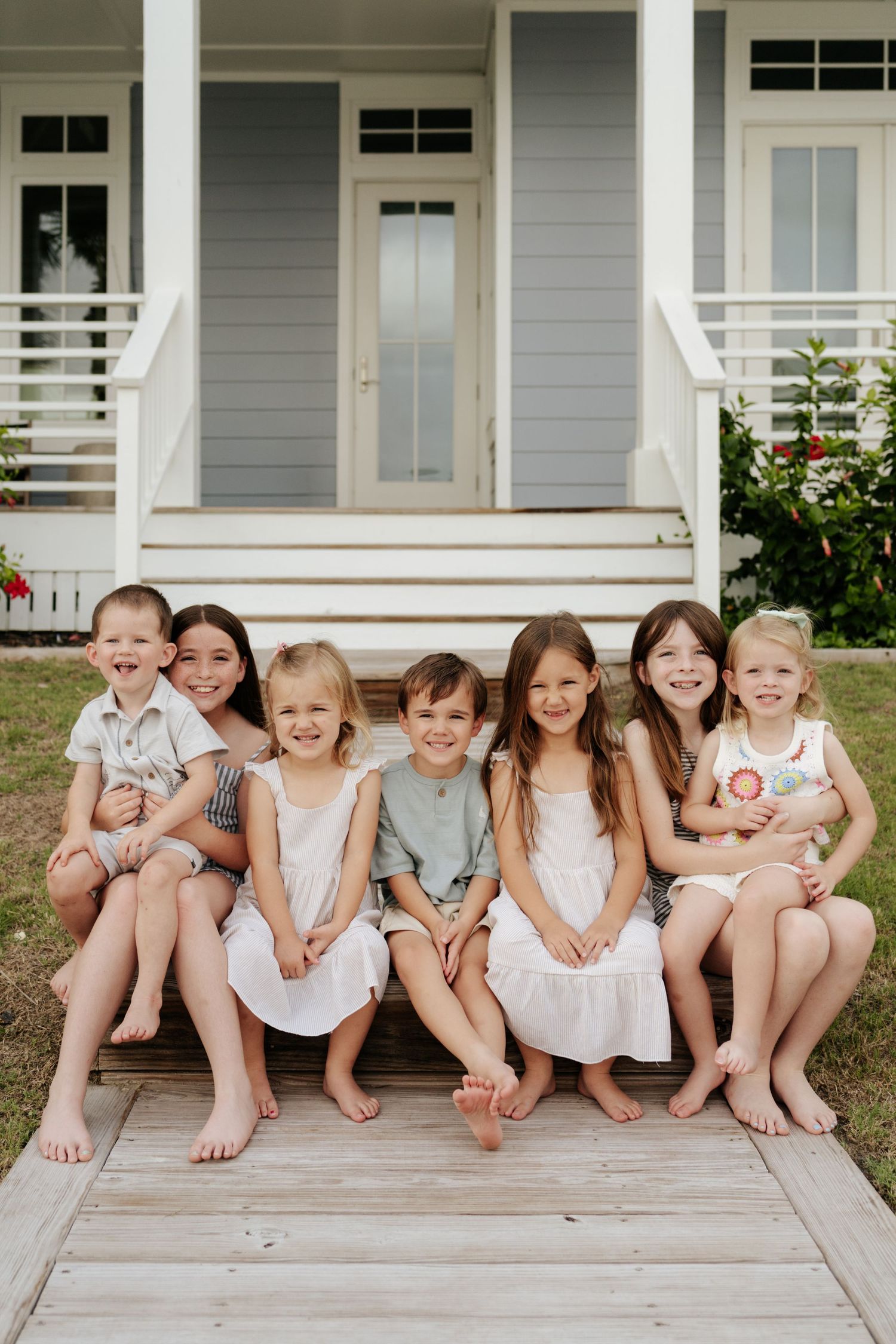 Seven smiling children sitting together on wooden porch steps in front of a gray house on a sunny day.