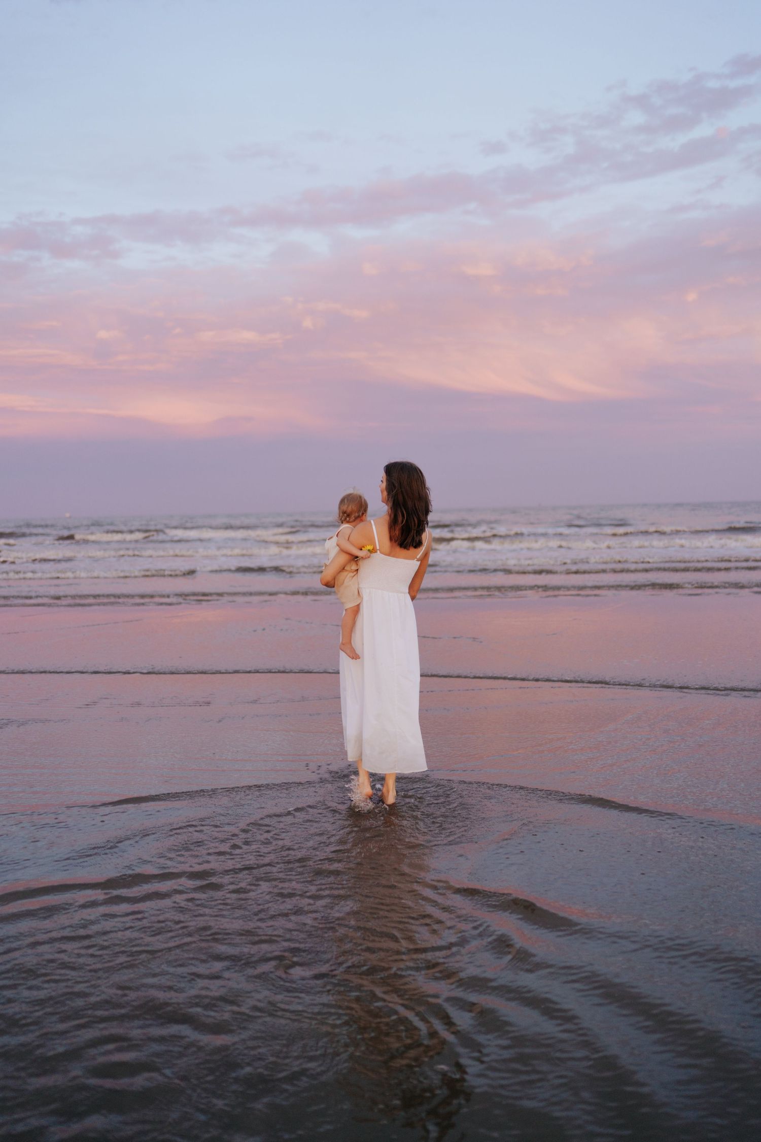 Mother in white dress holds baby at beach during pink sunset, standing in shallow ocean water.