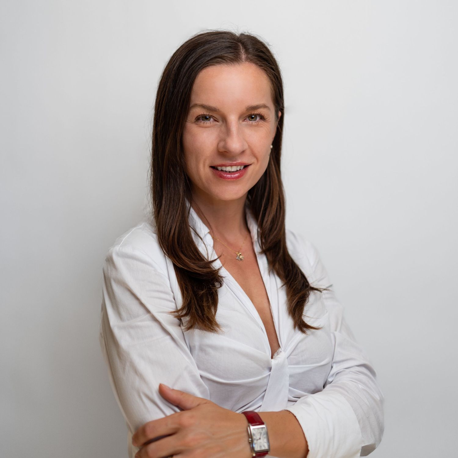 Professional woman with brown hair in white blouse, arms crossed, smiling confidently against neutral background.