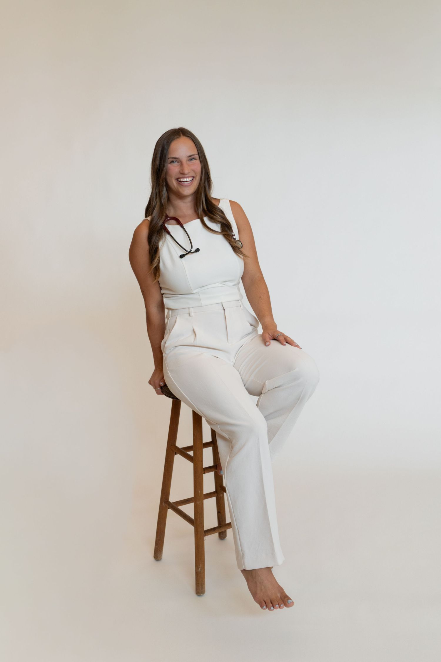 Smiling woman in white outfit with stethoscope sits on wooden stool against light background.