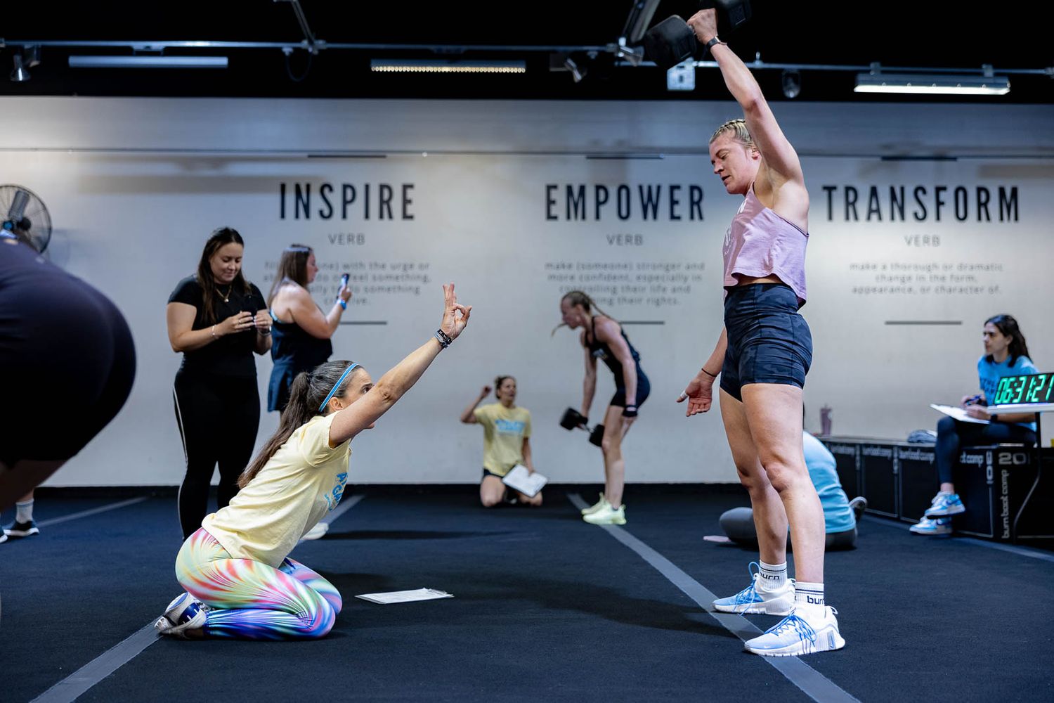 Two participants perform coordinated exercises beneath motivational wall text in a gym setting.