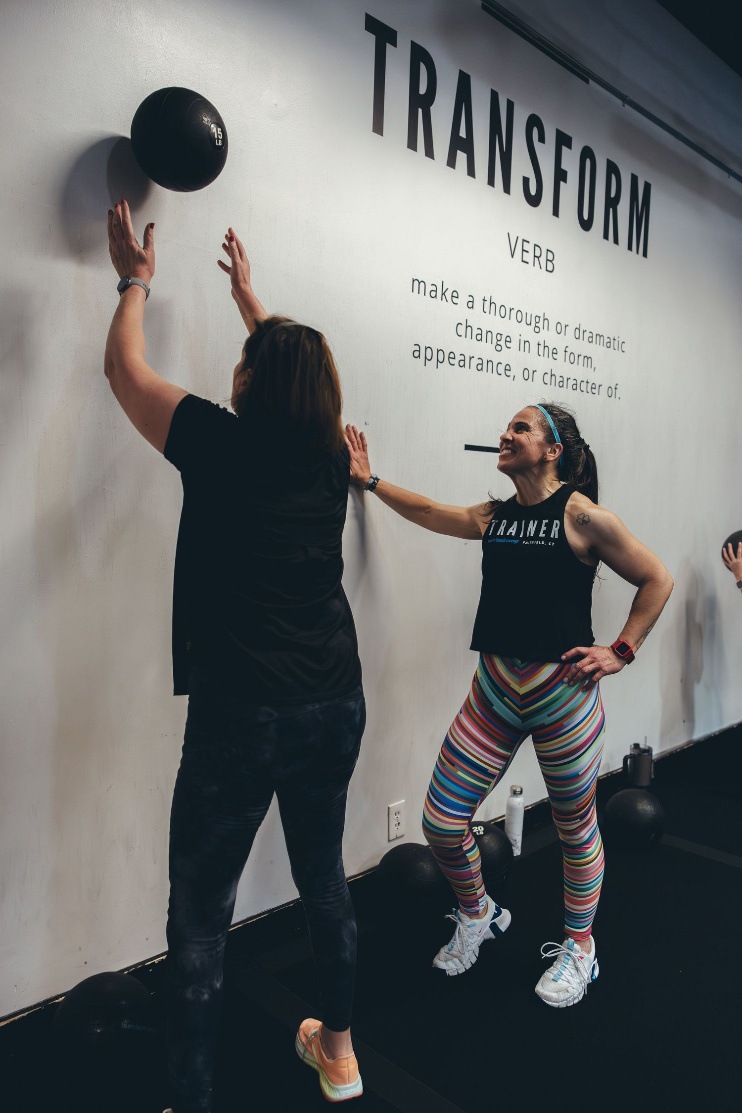 Two people engage in a medicine ball exercise beneath a 'Transform' wall text in a fitness studio.