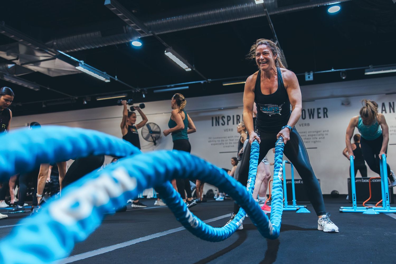 A group fitness class works out with blue battle ropes against a wall with motivational text.