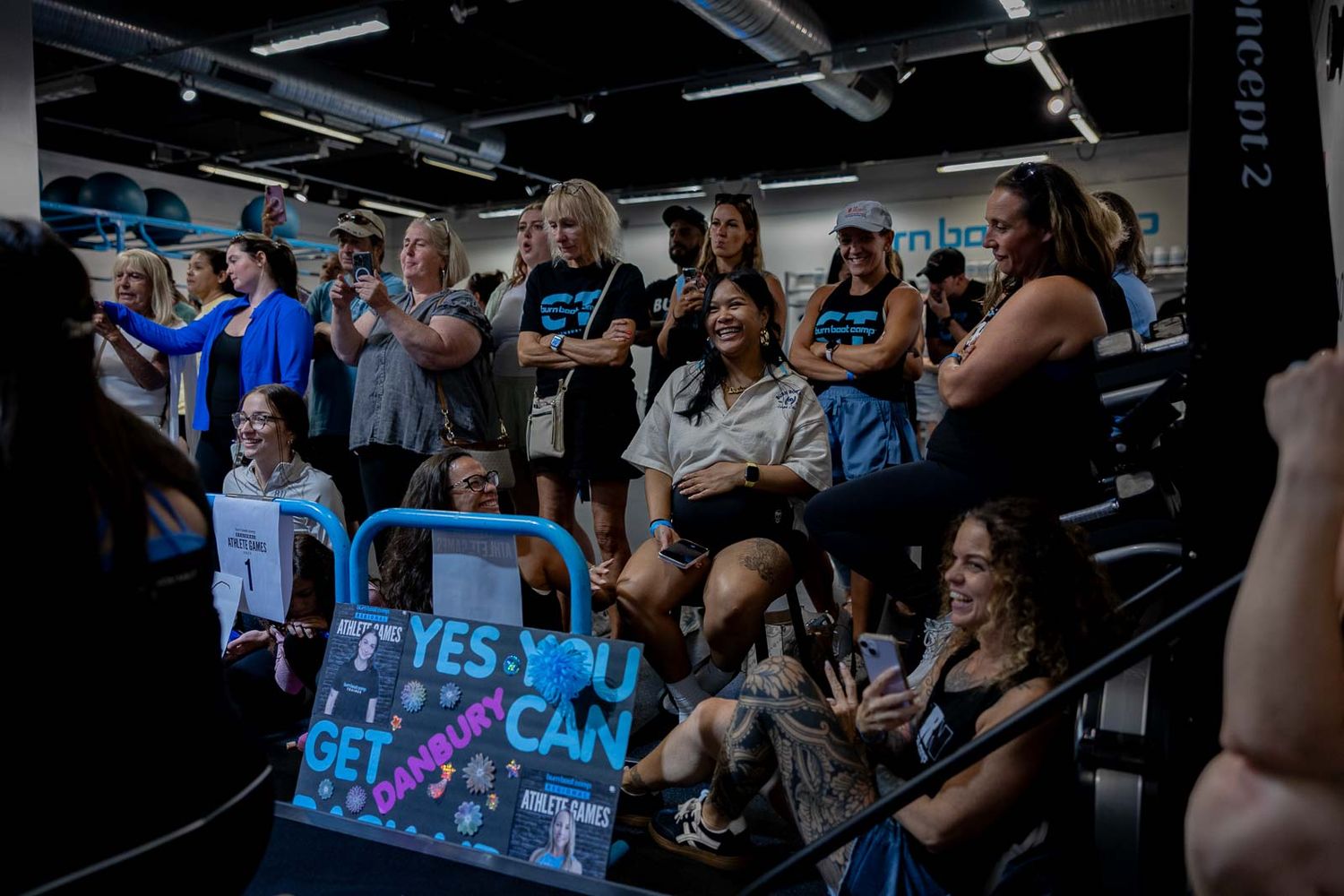 Fitness enthusiasts cheer and hold signs during a high-energy workout session at a gym.