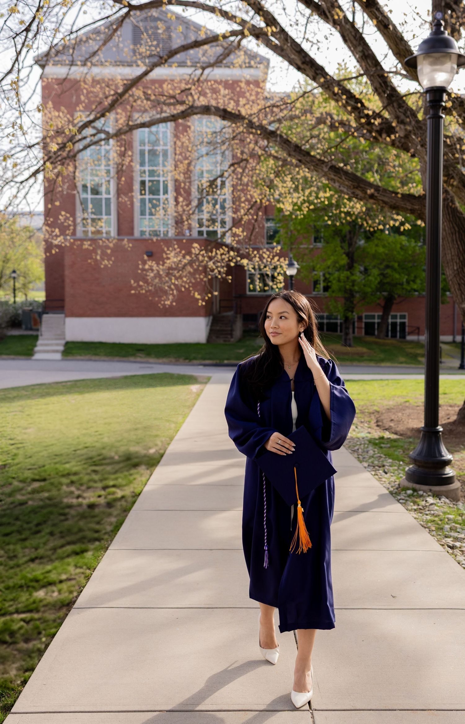 Graduate wearing navy blue graduation gown and cap walking along a campus sidewalk on a sunny spring day.