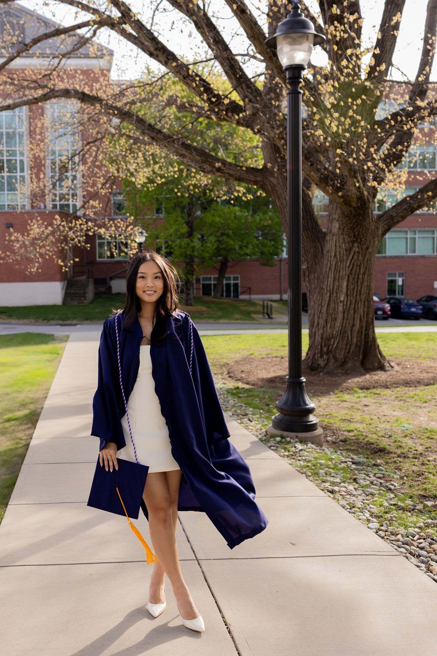 Graduate wearing navy graduation gown and white dress poses on campus sidewalk next to traditional lamp post.