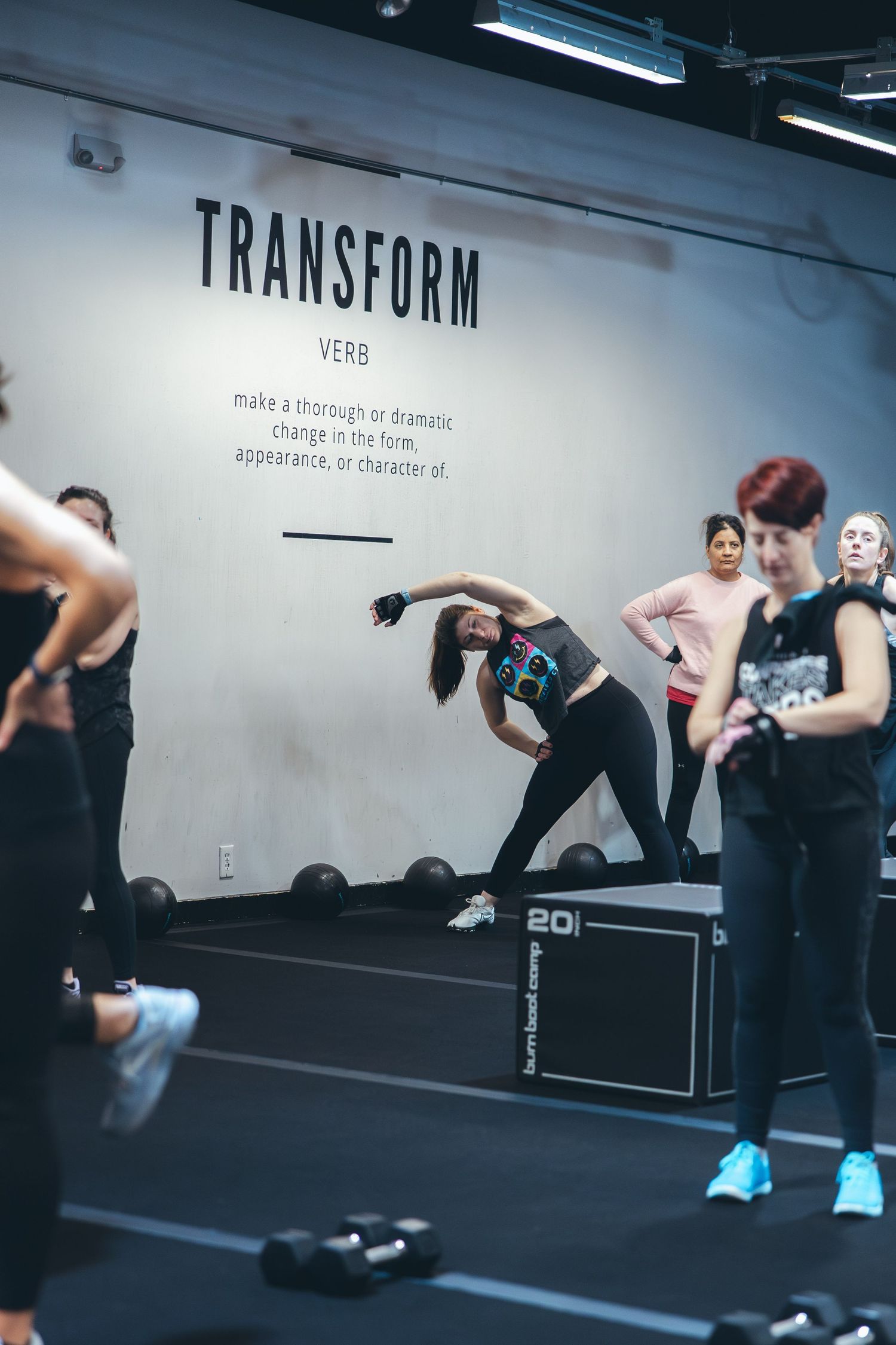Fitness class participants stretching and exercising in front of wall with 'Transform' text in modern gym studio.