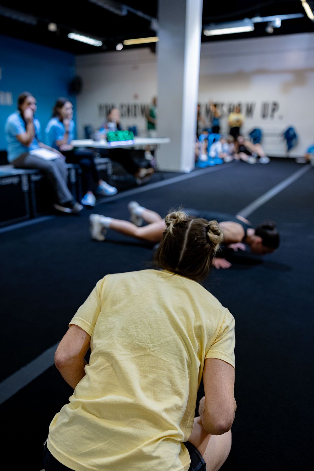Athletes exercising in a gym with dark flooring as others watch from tables in the background.