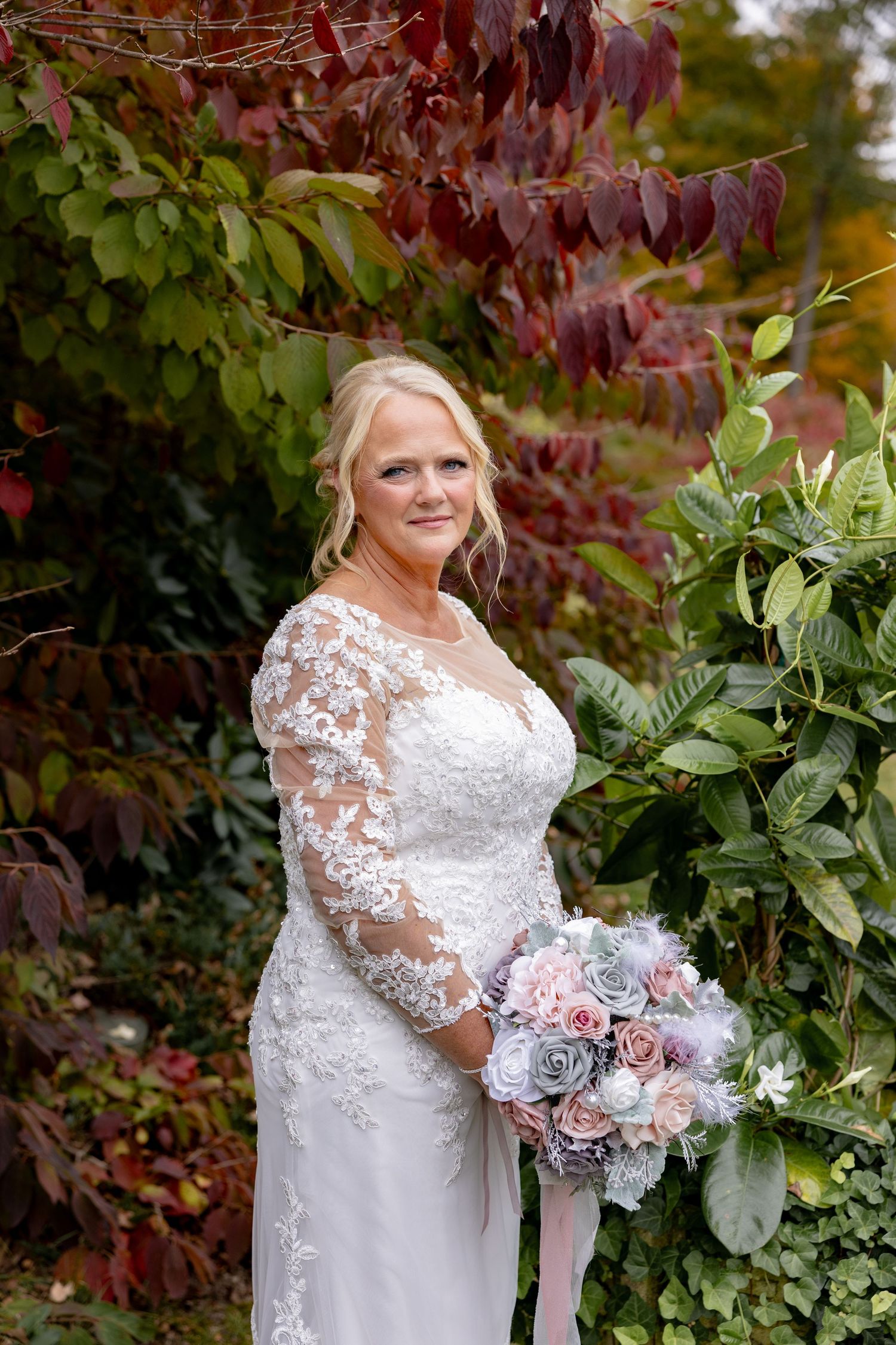 A bride in a white lace long-sleeve wedding dress holds a pink and white bouquet against autumn foliage.