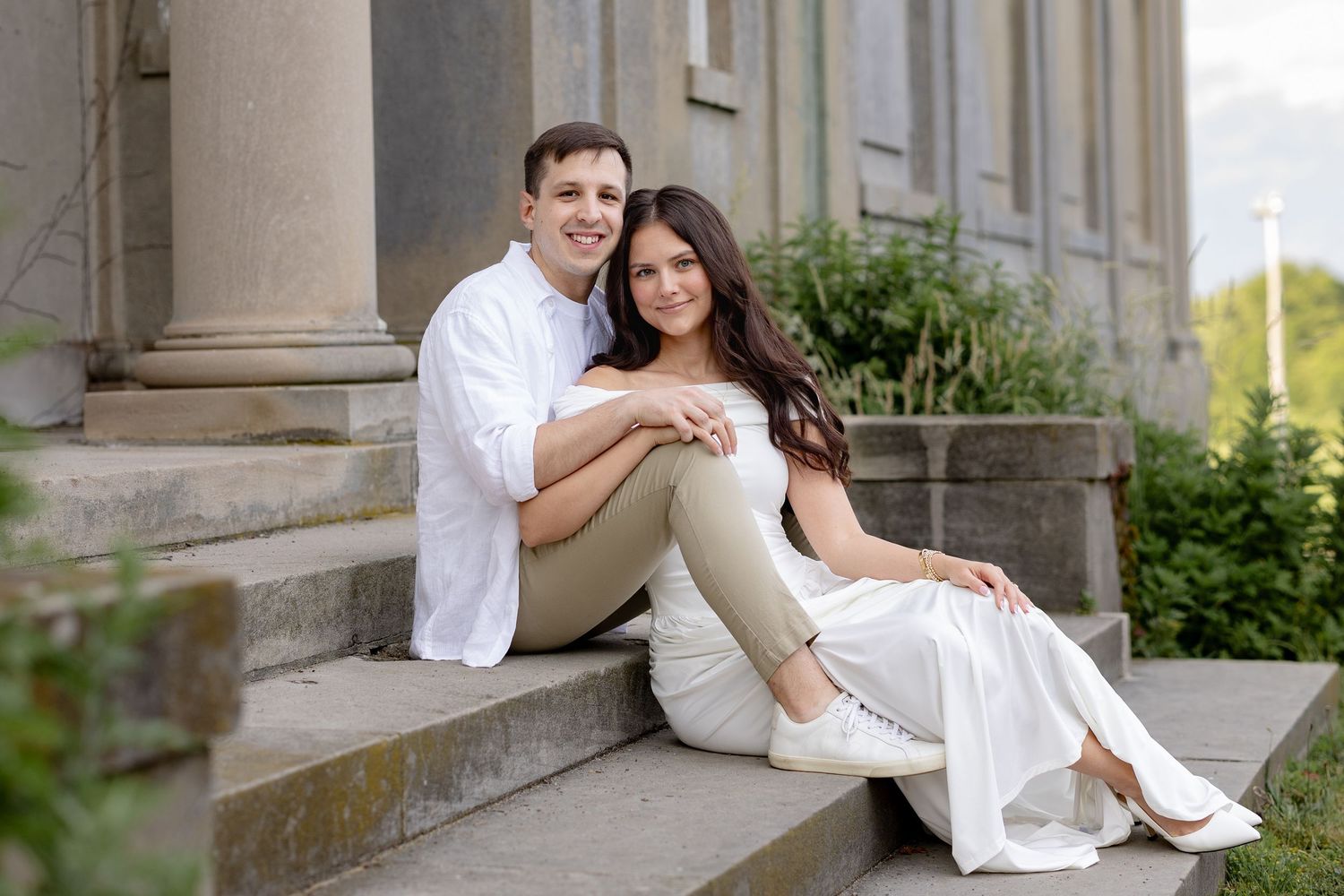 Elegant couple in white attire pose romantically on stone steps of historic building during wedding photo session.