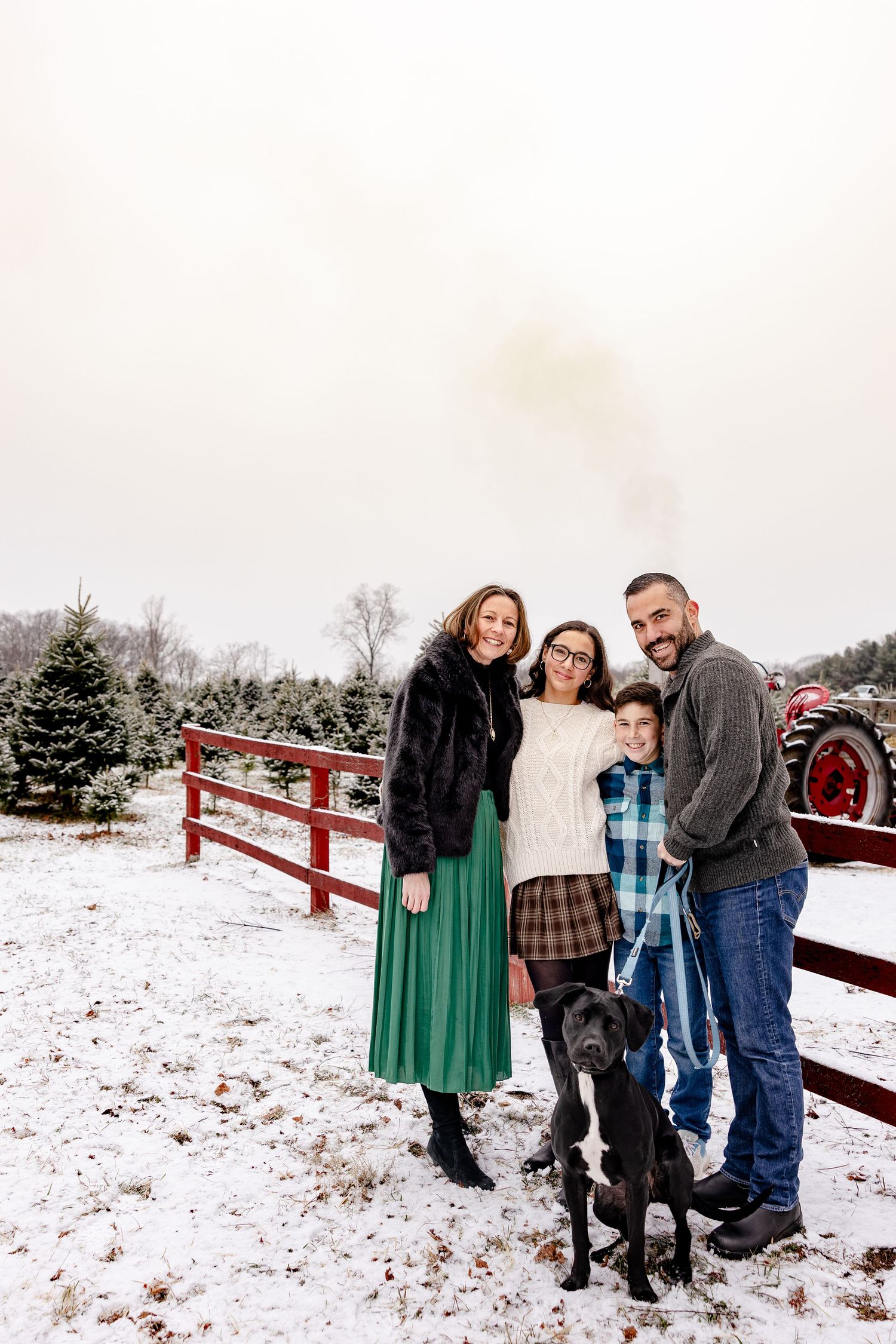 Family and dog pose together in snowy winter scene at Christmas tree farm with red fence and evergreens in background.