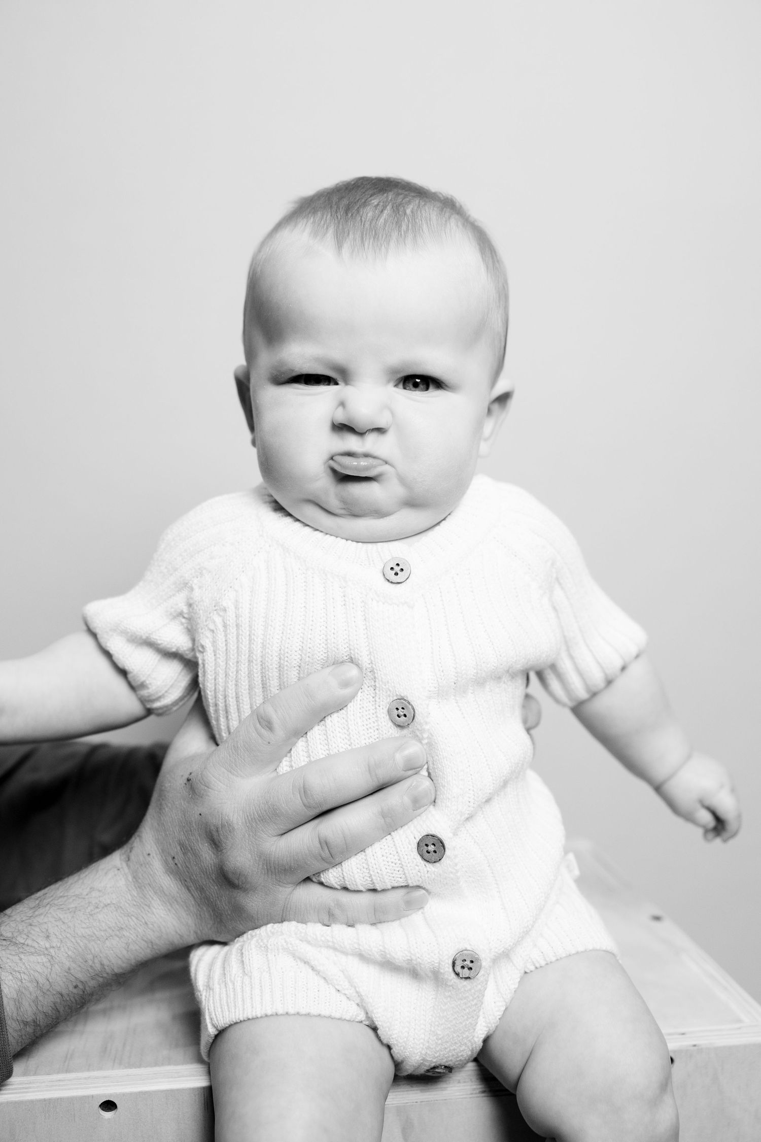 Black and white image of a baby in knit romper making pouty expression while supported by hands.