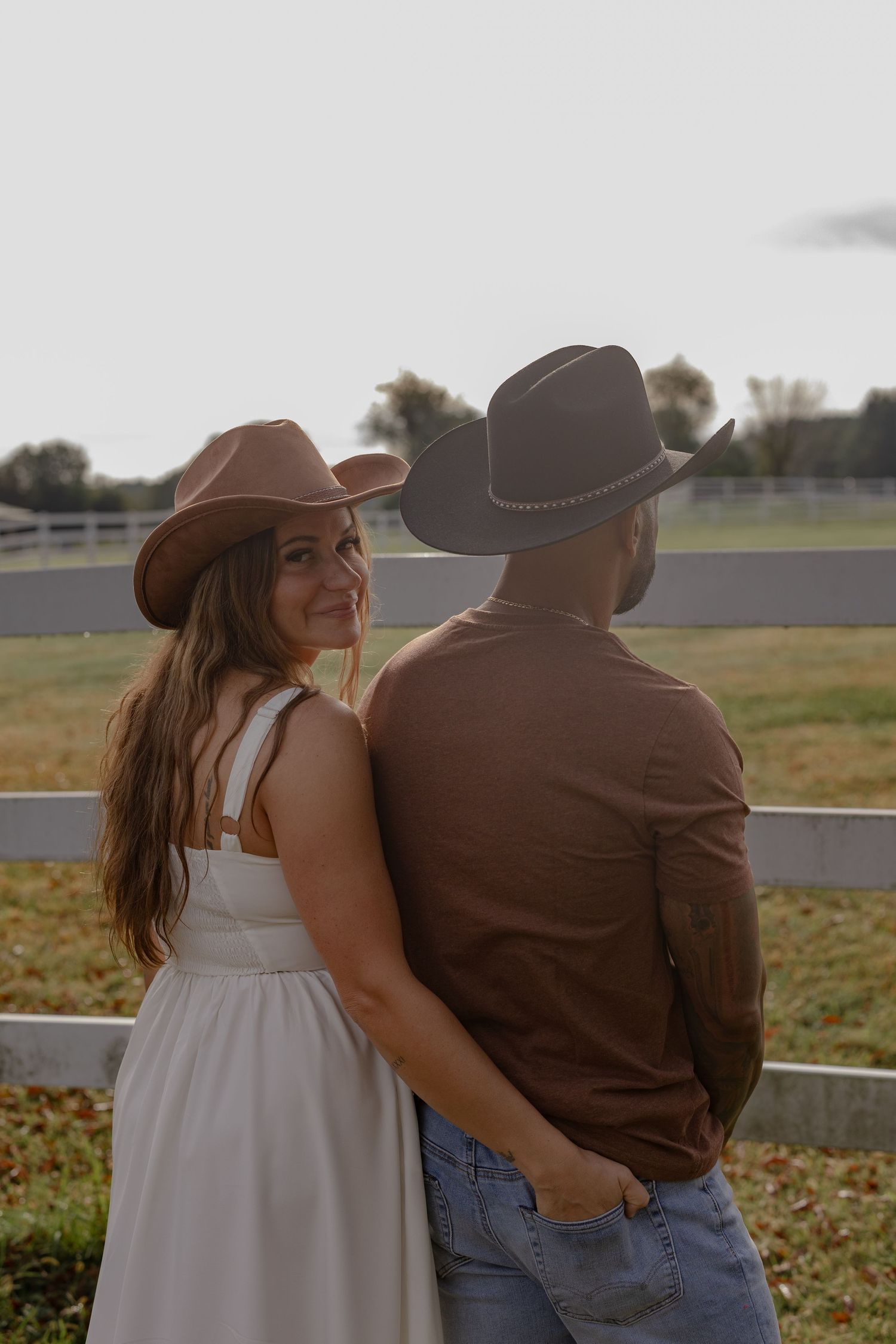 A couple in casual attire stands together by a rustic fence, looking out over a rural field on an autumn day.