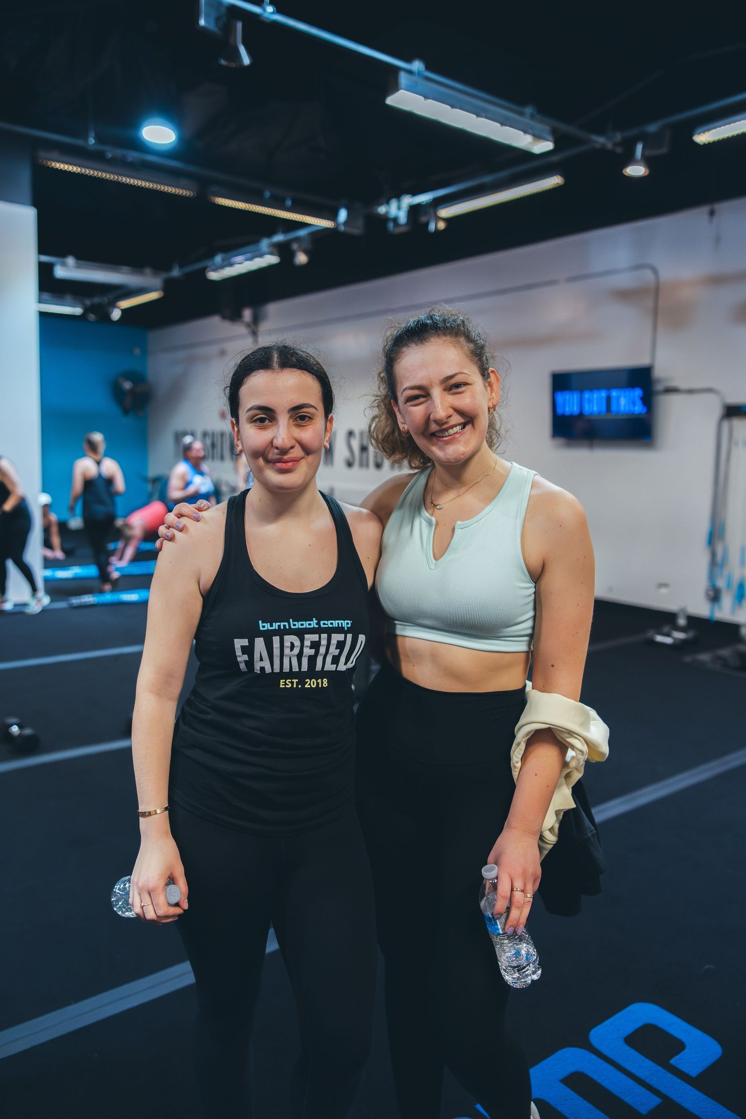 Two workout partners smiling and holding water bottles in a modern fitness gym with blue walls.