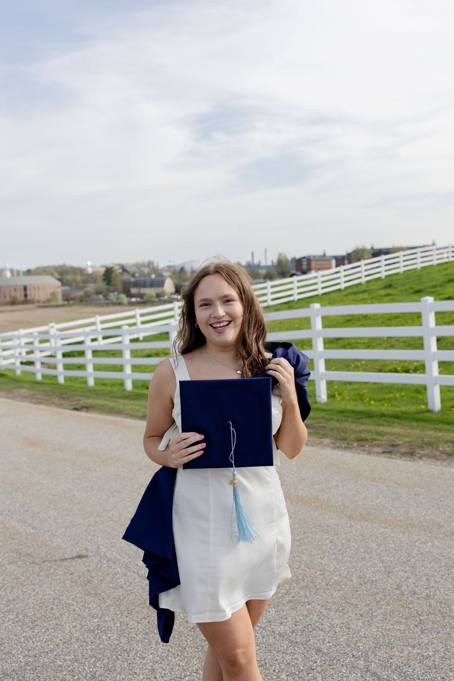 Graduate in white dress with navy blue graduation gown and diploma celebrates outdoors by white fence at countryside.