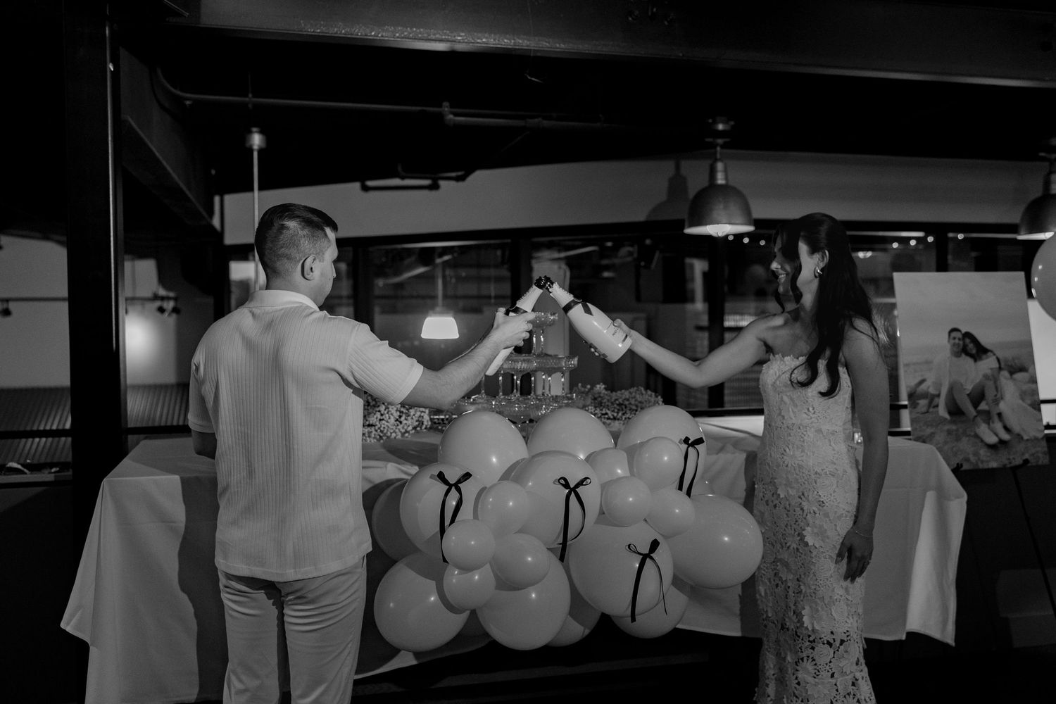 A couple toasts with champagne glasses in front of a decorated balloon display at a celebratory event.