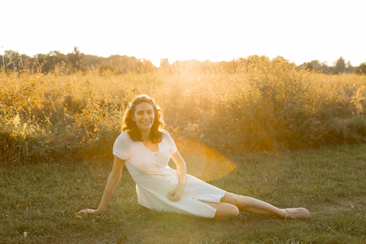 A figure in a white dress lays gracefully in a sunlit meadow during golden hour.